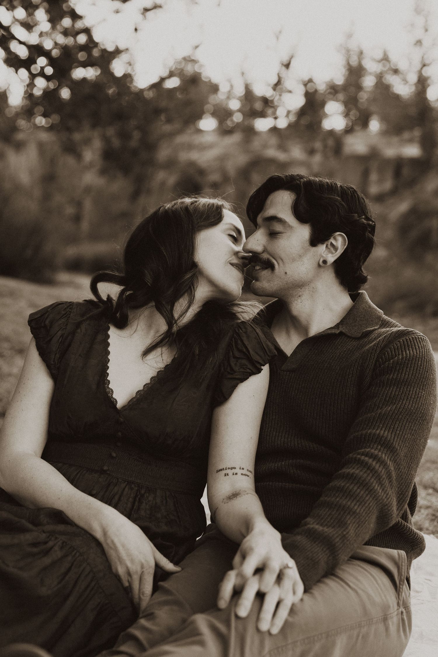 Romantic couple sharing a kiss outdoors in a sepia-toned portrait, sitting together in a natural setting.