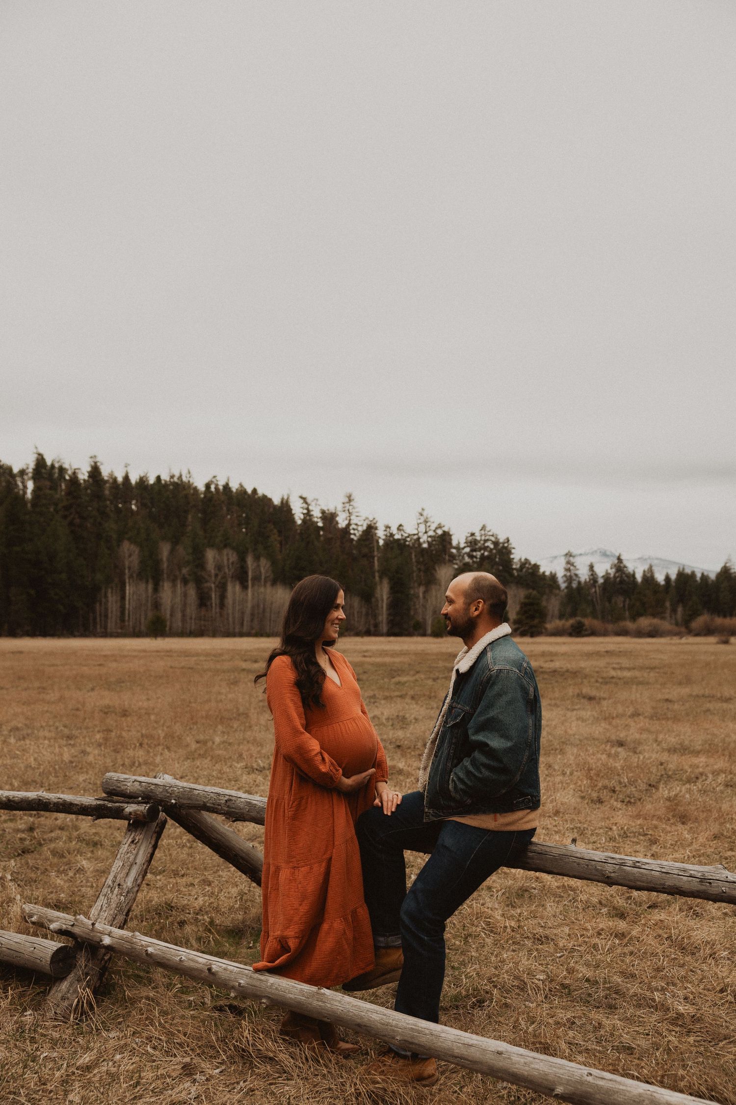 Pregnant woman in rust orange dress holds hands with partner by a wooden fence in an autumn field.