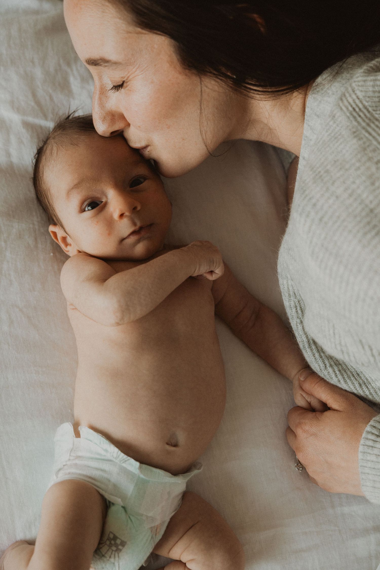 A mother tenderly kisses her newborn baby lying on a white blanket, sharing a peaceful bonding moment.