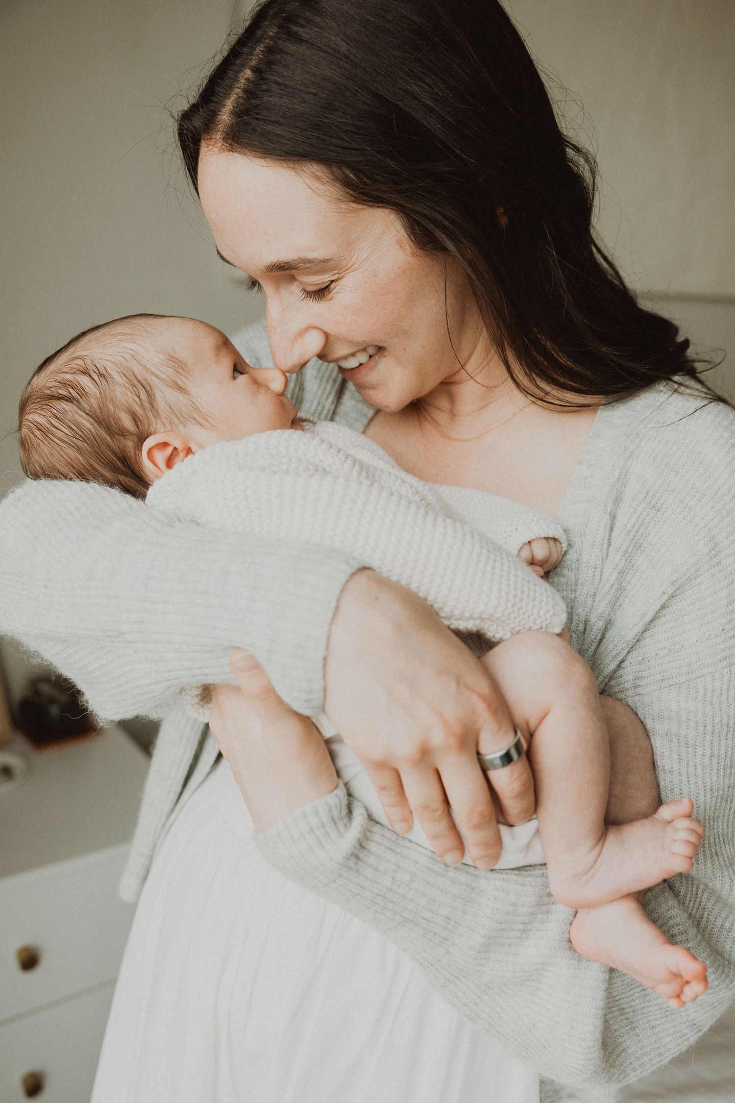A smiling mother with dark hair tenderly holds her newborn baby close, touching noses in a bright nursery room.