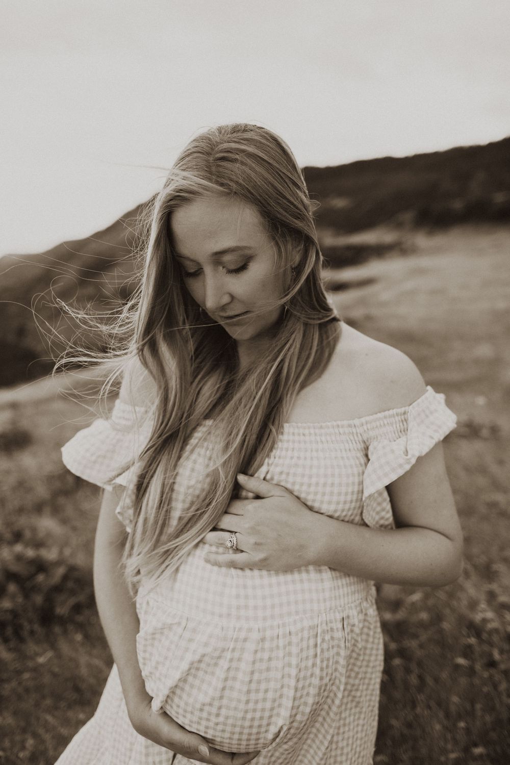 Woman in a white gingham dress poses thoughtfully in a windswept field.