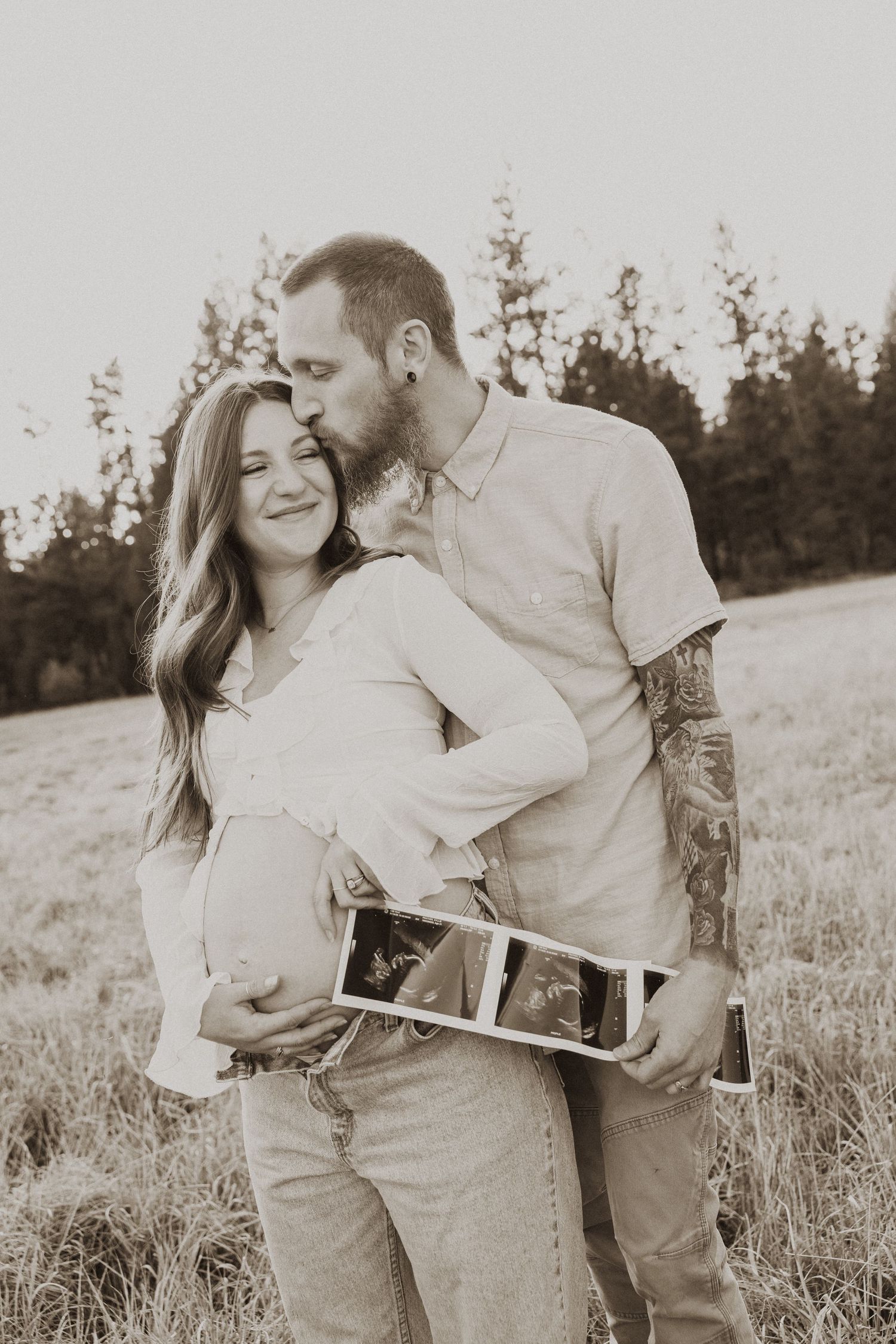 Couple in sepia-toned maternity photo outdoors, man kissing woman's head while she holds ultrasound images in a field.