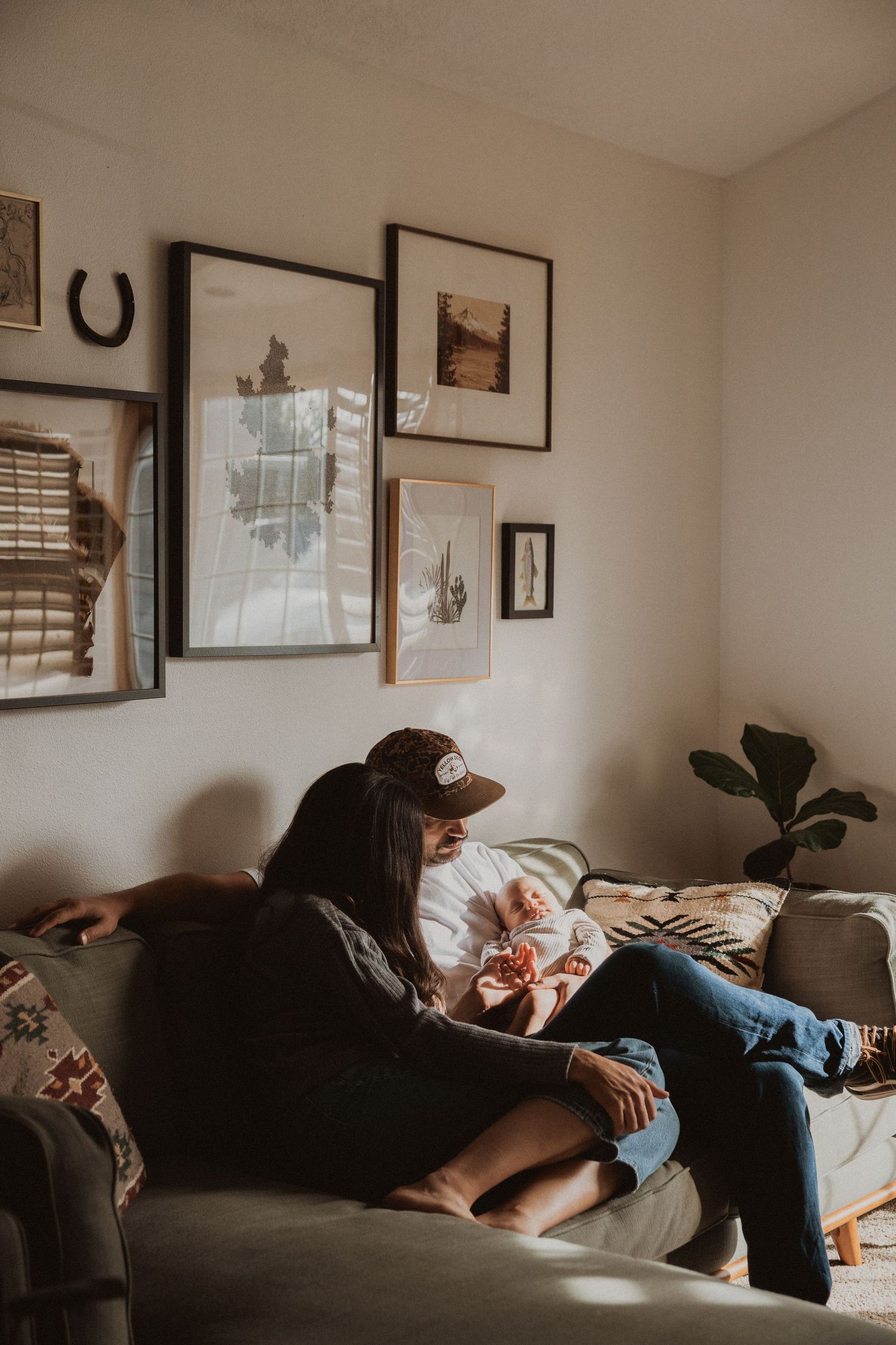Couple sitting on couch holding newborn baby in cozy living room with gallery wall and natural light.