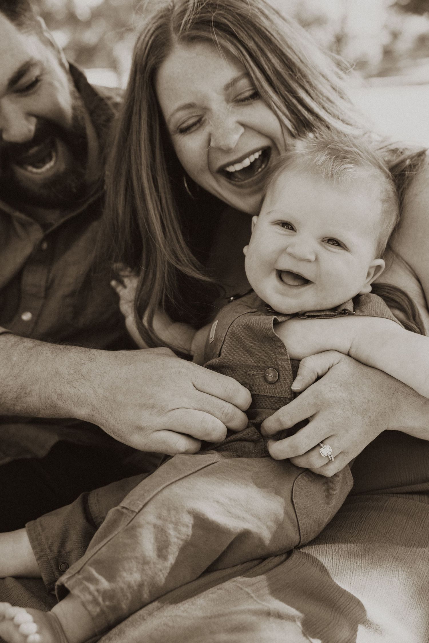 Sepia-toned photo of laughing parents holding their smiling baby during a joyful family portrait session.