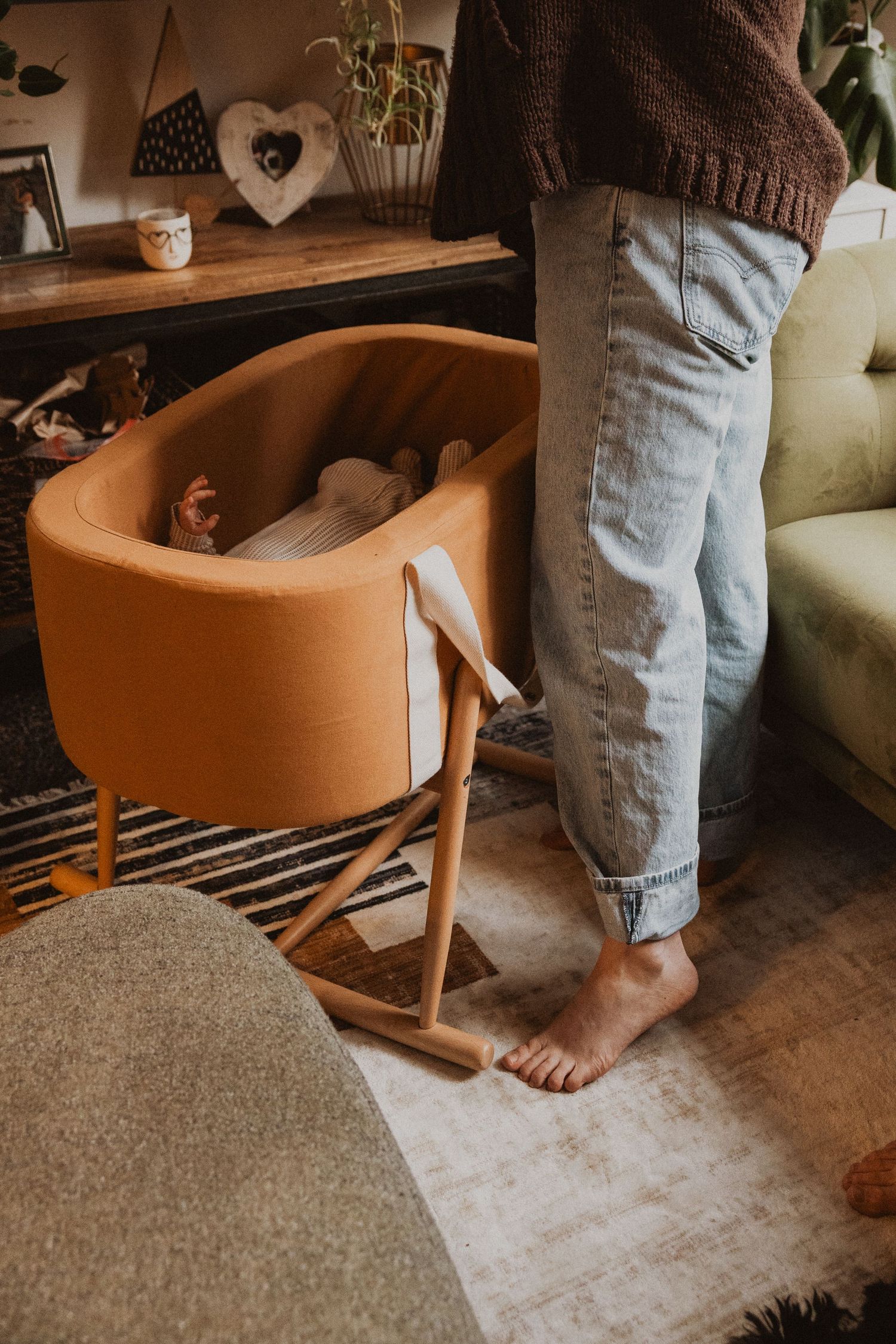 Parent standing barefoot beside a modern mustard yellow wooden bassinet with sleeping newborn.