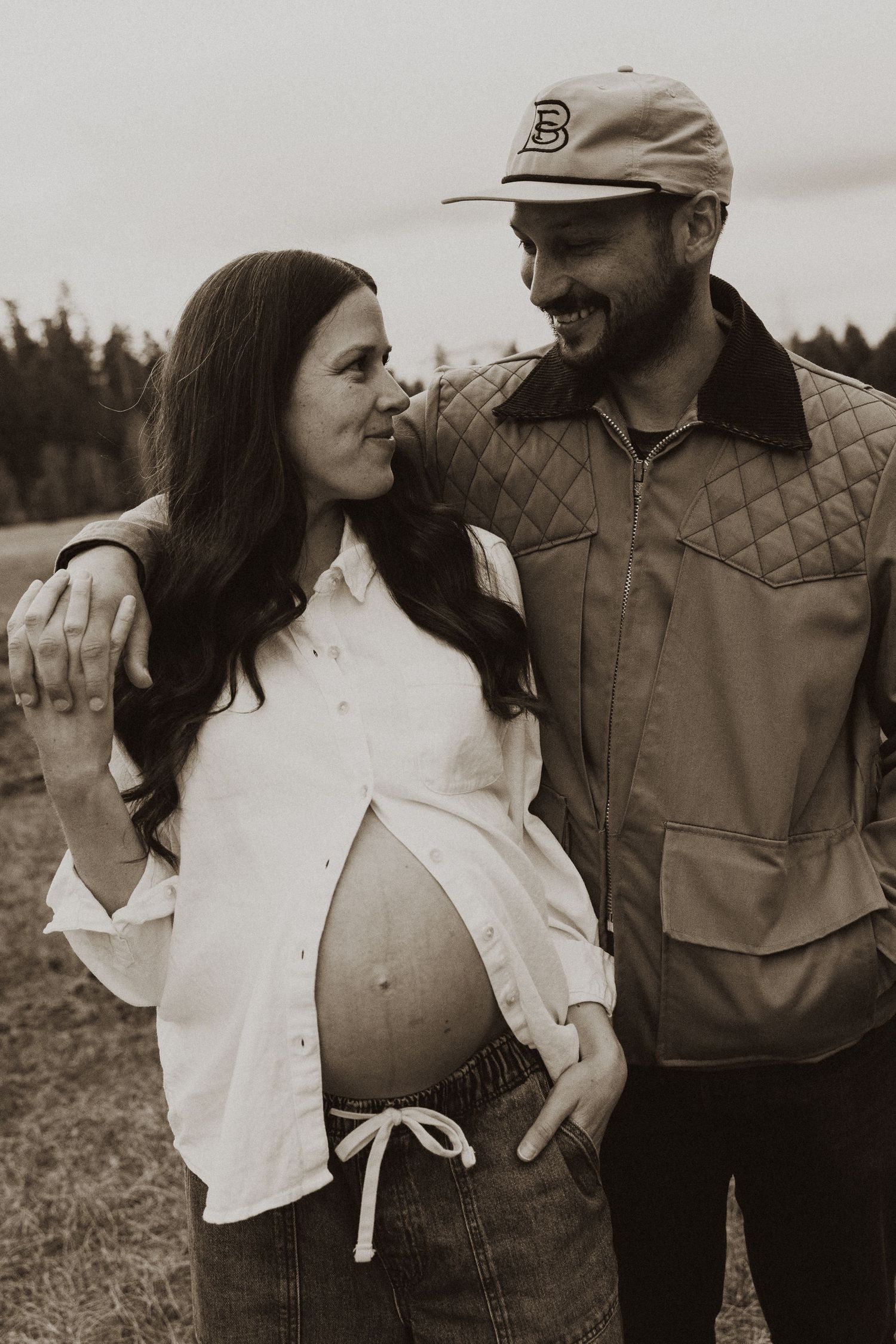 Sepia photo of smiling pregnant couple standing together in an open field, man wearing a cap.