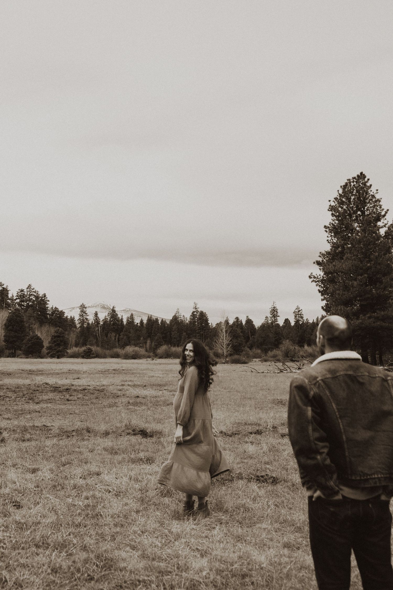Pregnant woman in long dress smiles back at her partner while standing in a wide open meadow.