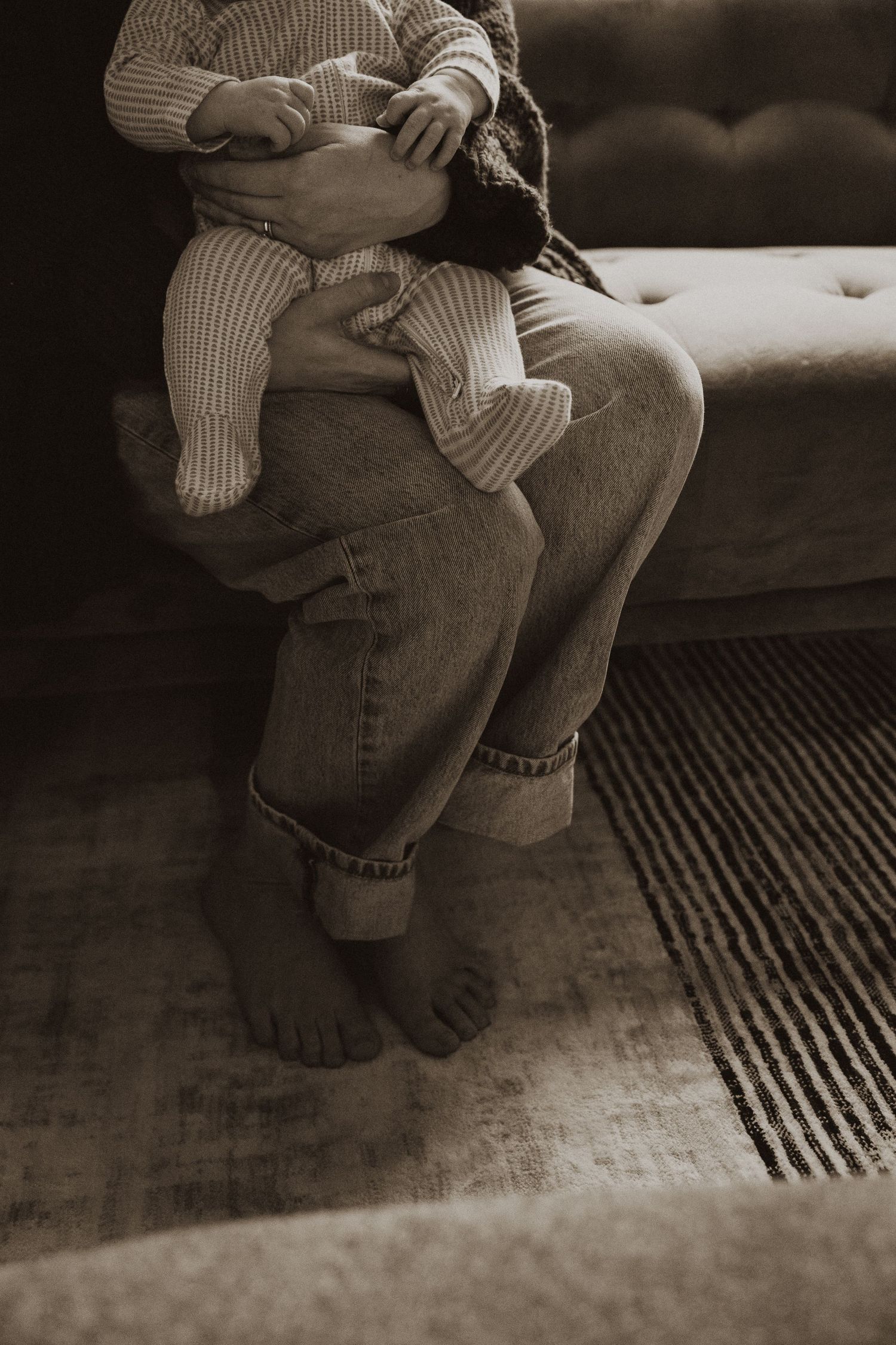 Parent sitting on couch holding newborn baby in striped onesie in sepia-toned portrait.