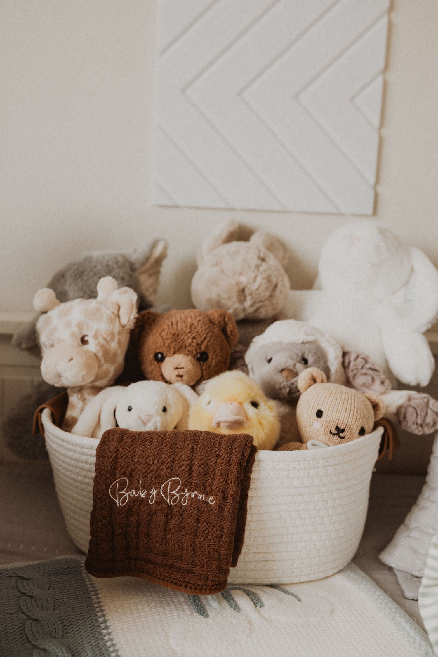 White rope basket filled with stuffed animals and a brown knit blanket labeled Baby Byrne in a cozy nursery setting.
