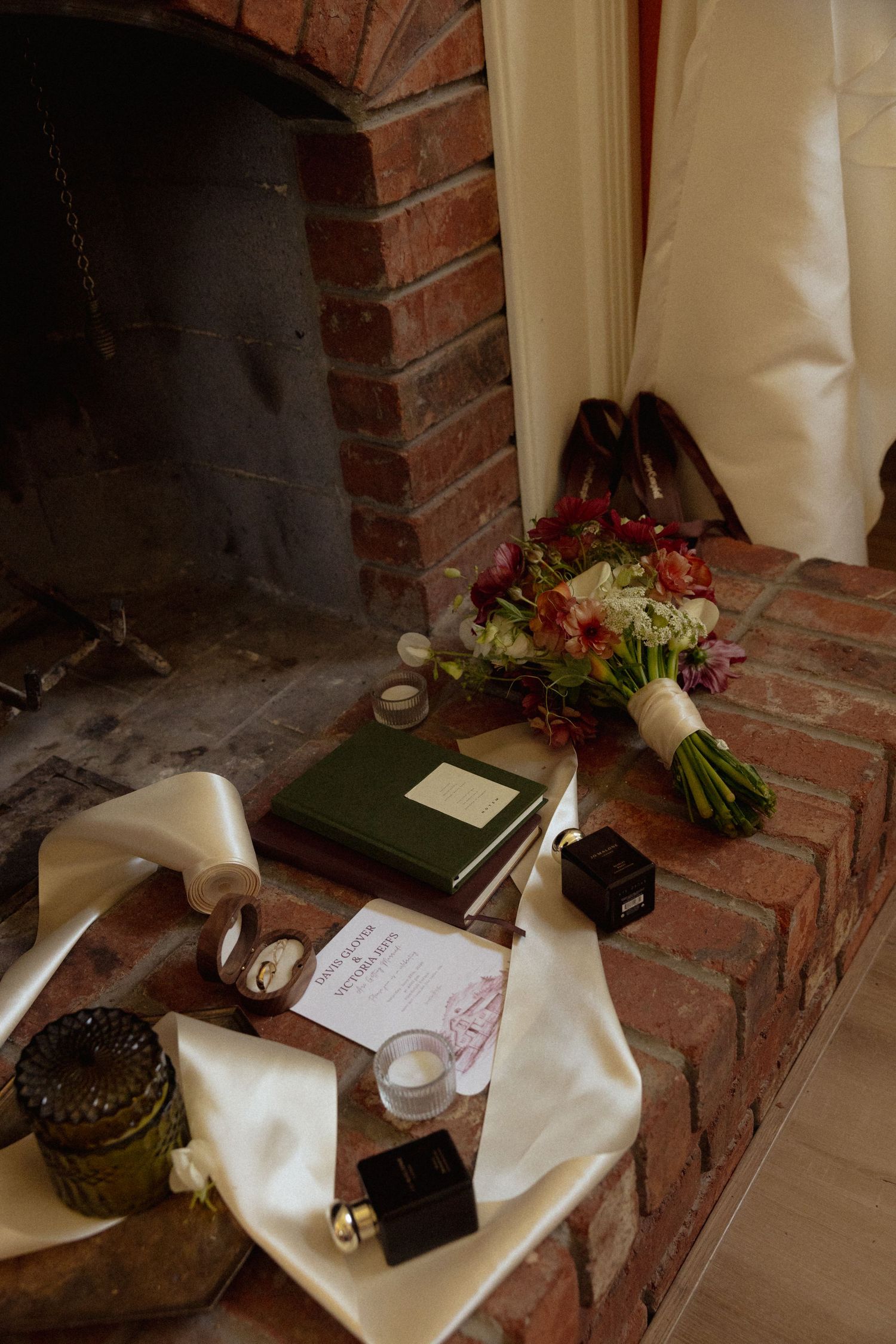 Wedding flat lay with floral bouquet, green journal, invitation, and perfume on a rustic brick fireplace hearth.