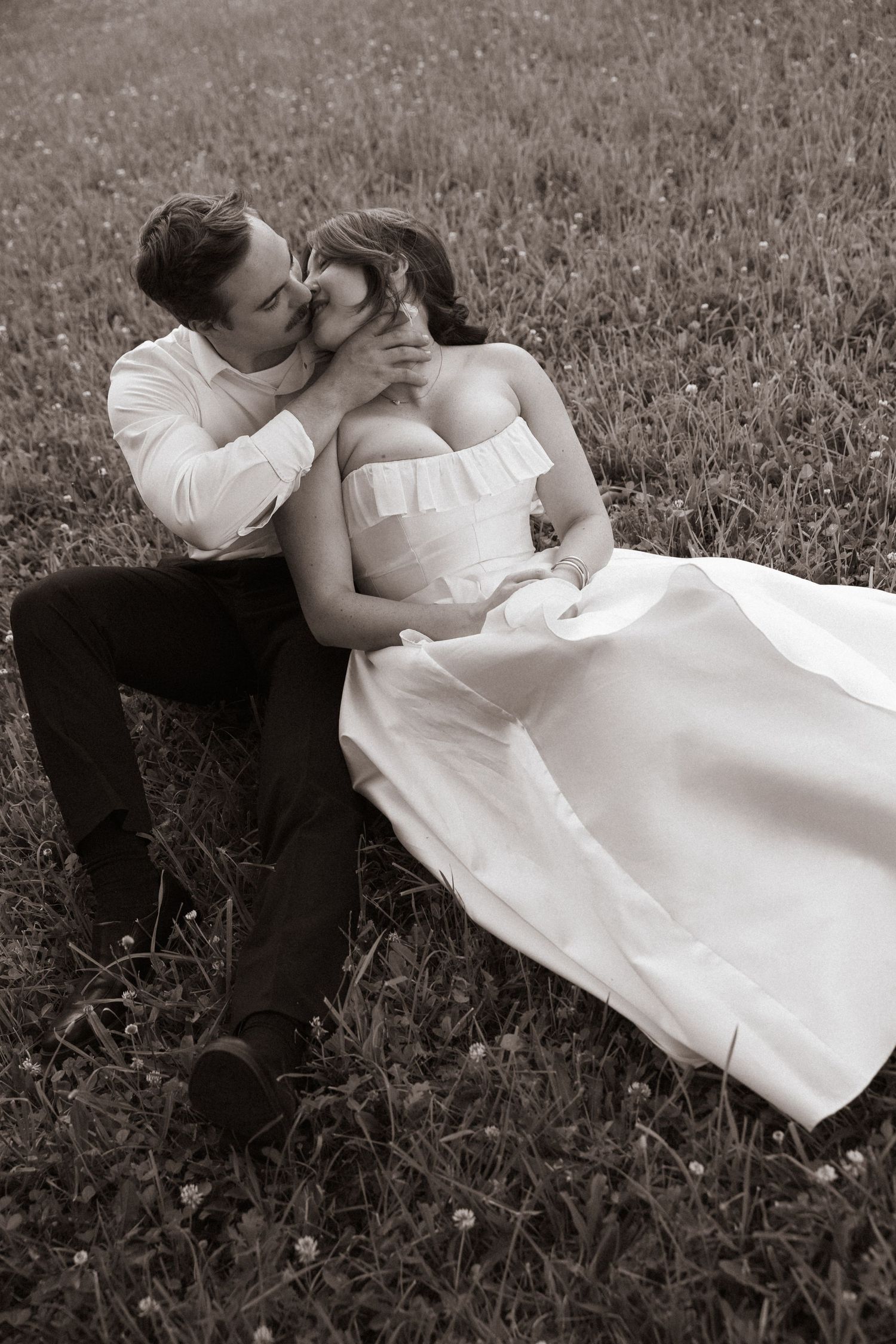 Romantic black and white photo of groom tenderly kissing bride while sitting together in a wildflower meadow.