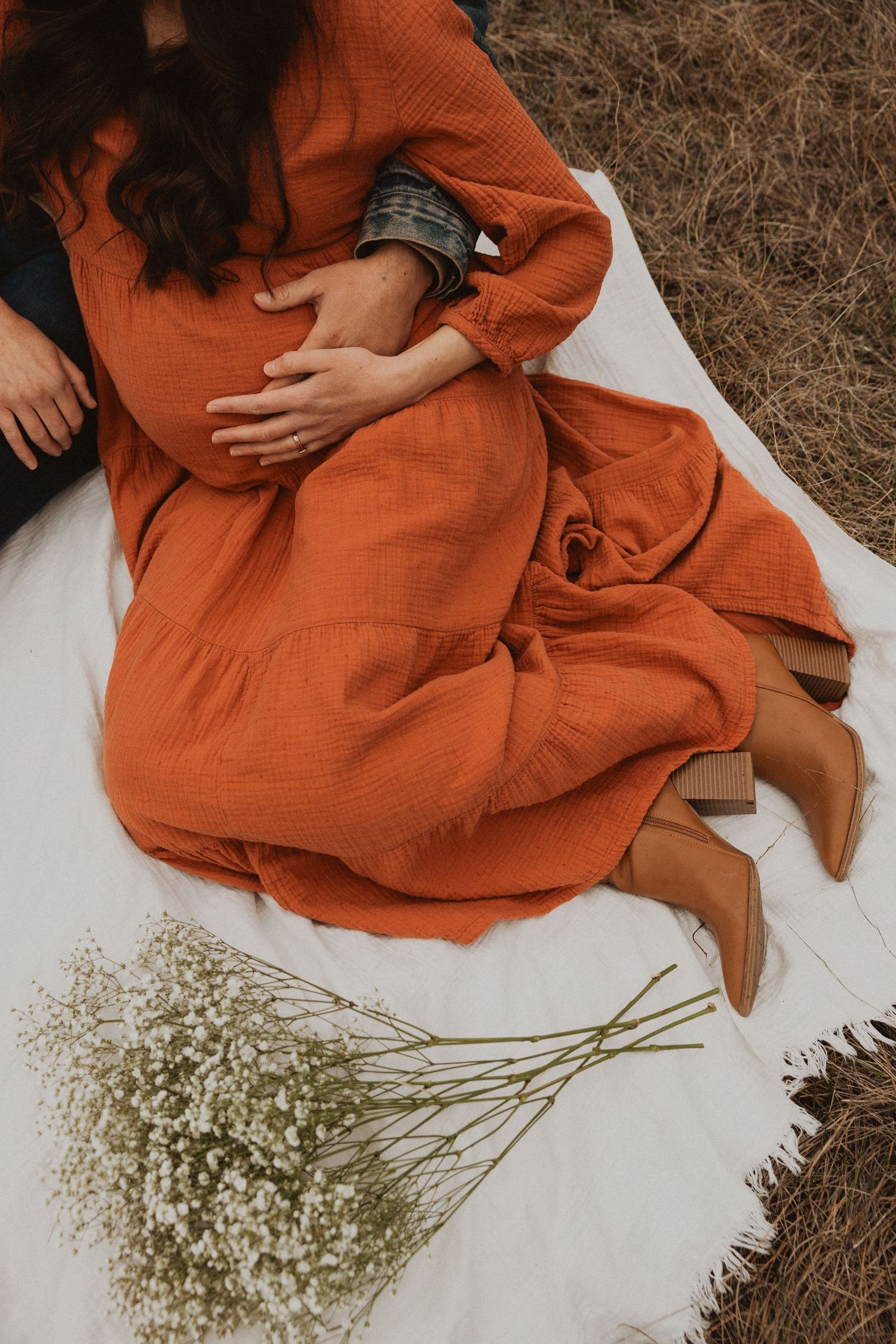 Close-up of pregnant woman in rust orange dress sitting on white blanket with baby's breath flowers nearby.