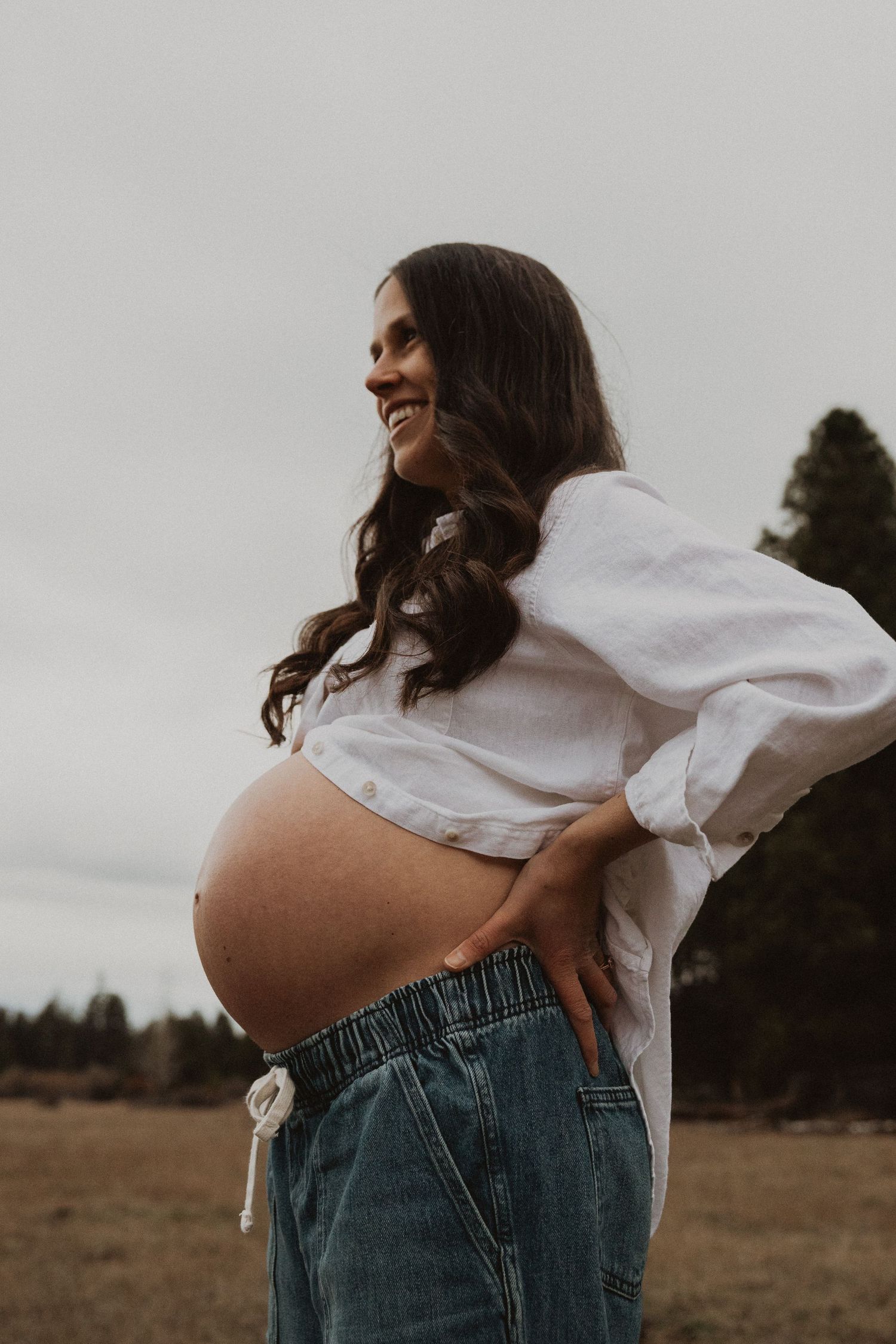 Smiling pregnant woman in white top and jeans poses outdoors in a field with trees in background.