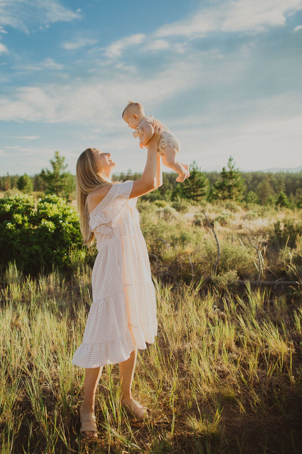 Joyful outdoor portrait of parent lifting baby against blue sky and pine trees.