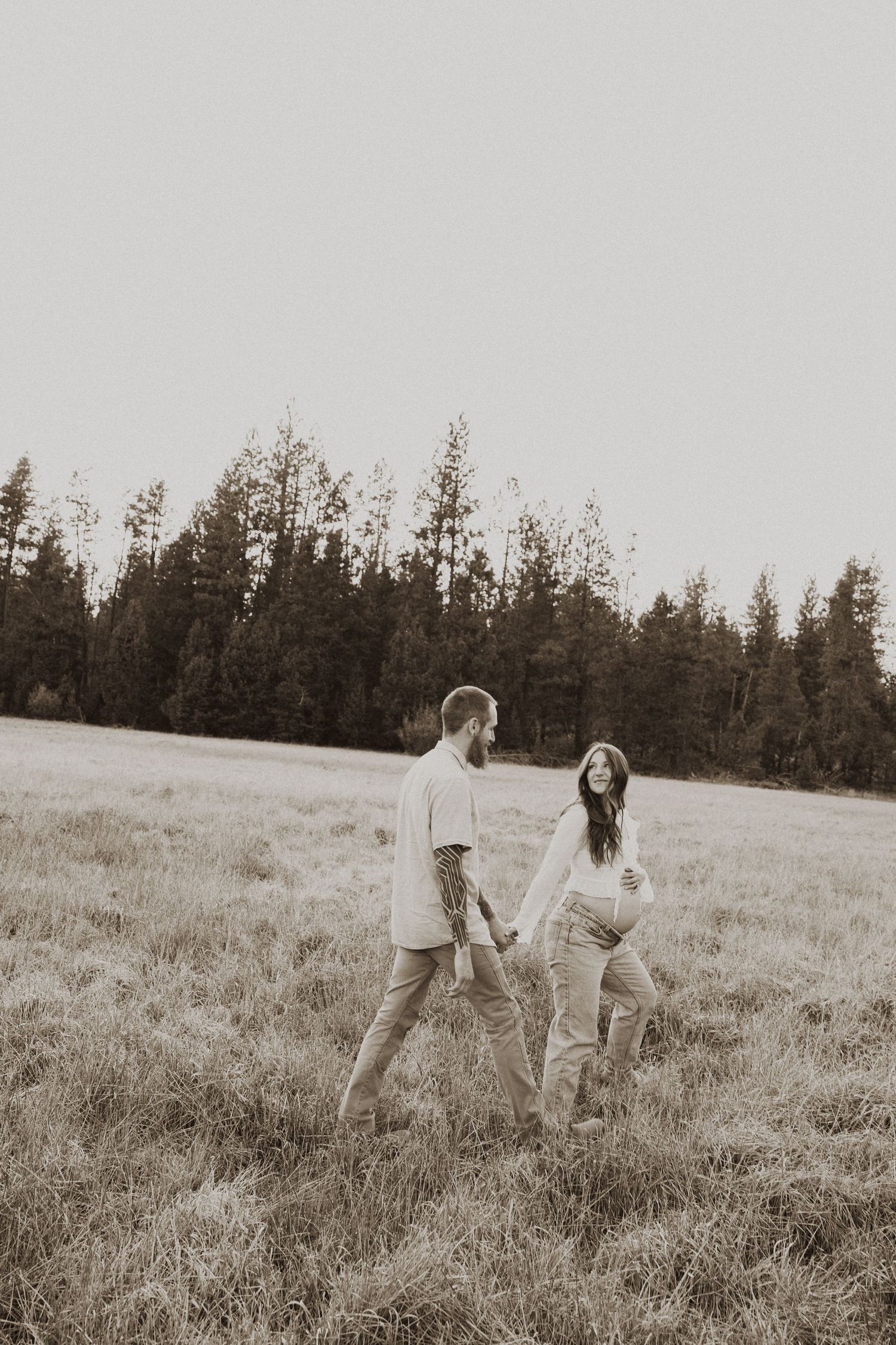 A happy expectant couple holds hands while walking through a grassy meadow surrounded by tall pine trees.