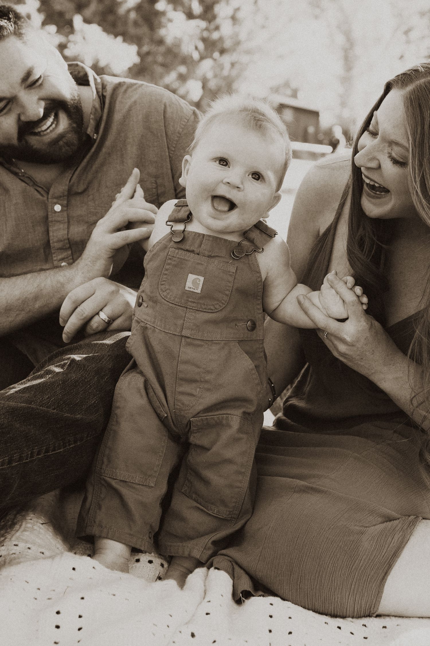 Sepia photo of happy parents laughing with their baby in overalls during a family portrait session outdoors.