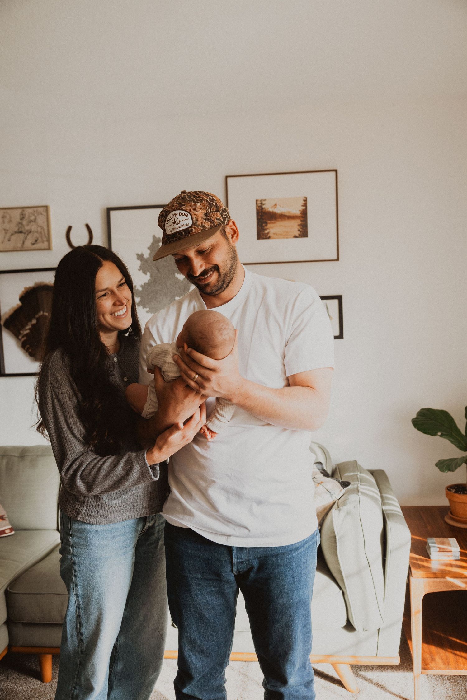 Happy couple smiling at their newborn baby in a cozy living room with framed art on the wall.