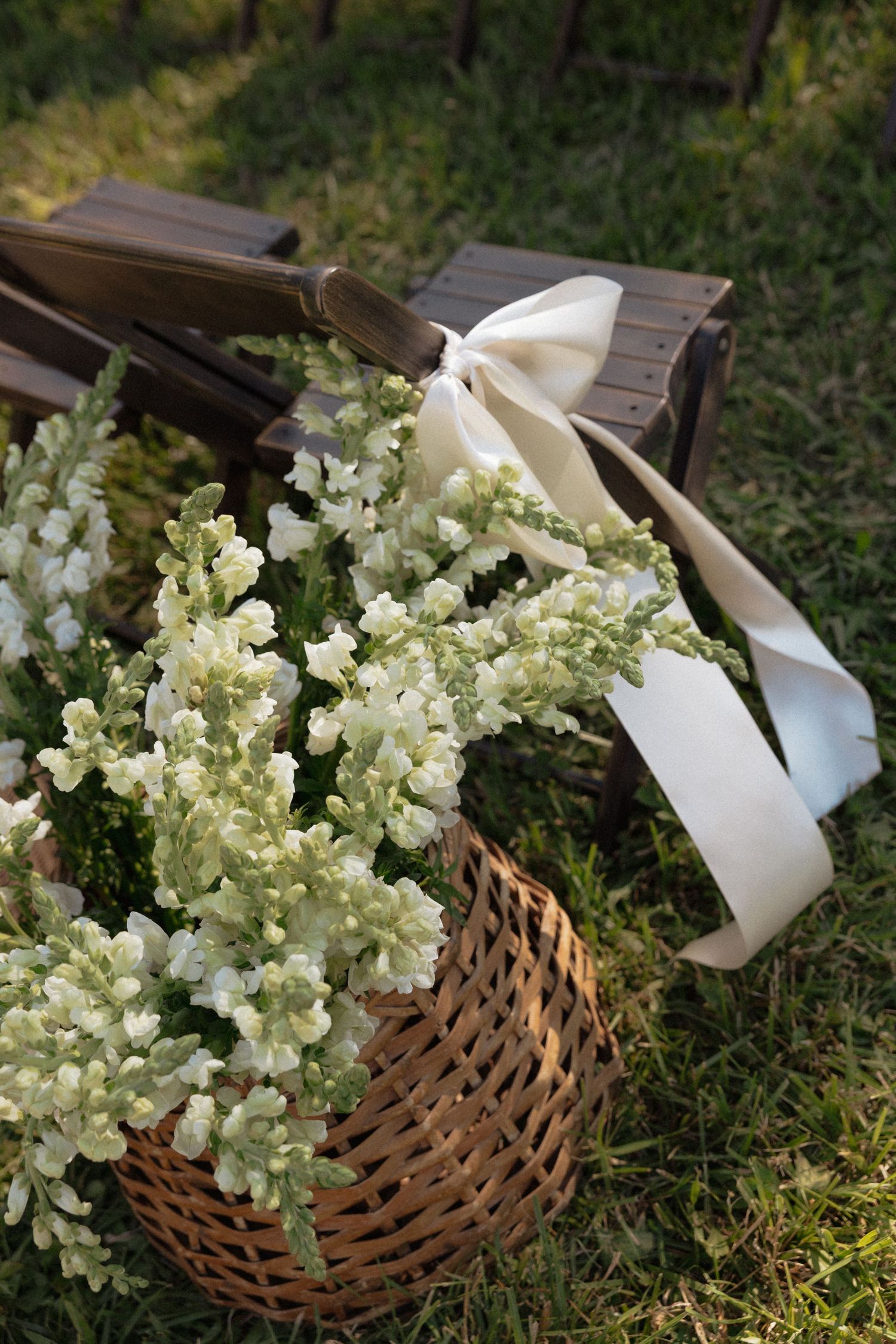 Wicker basket filled with white snapdragon flowers tied with a satin bow rests beside a wooden bench on grass.