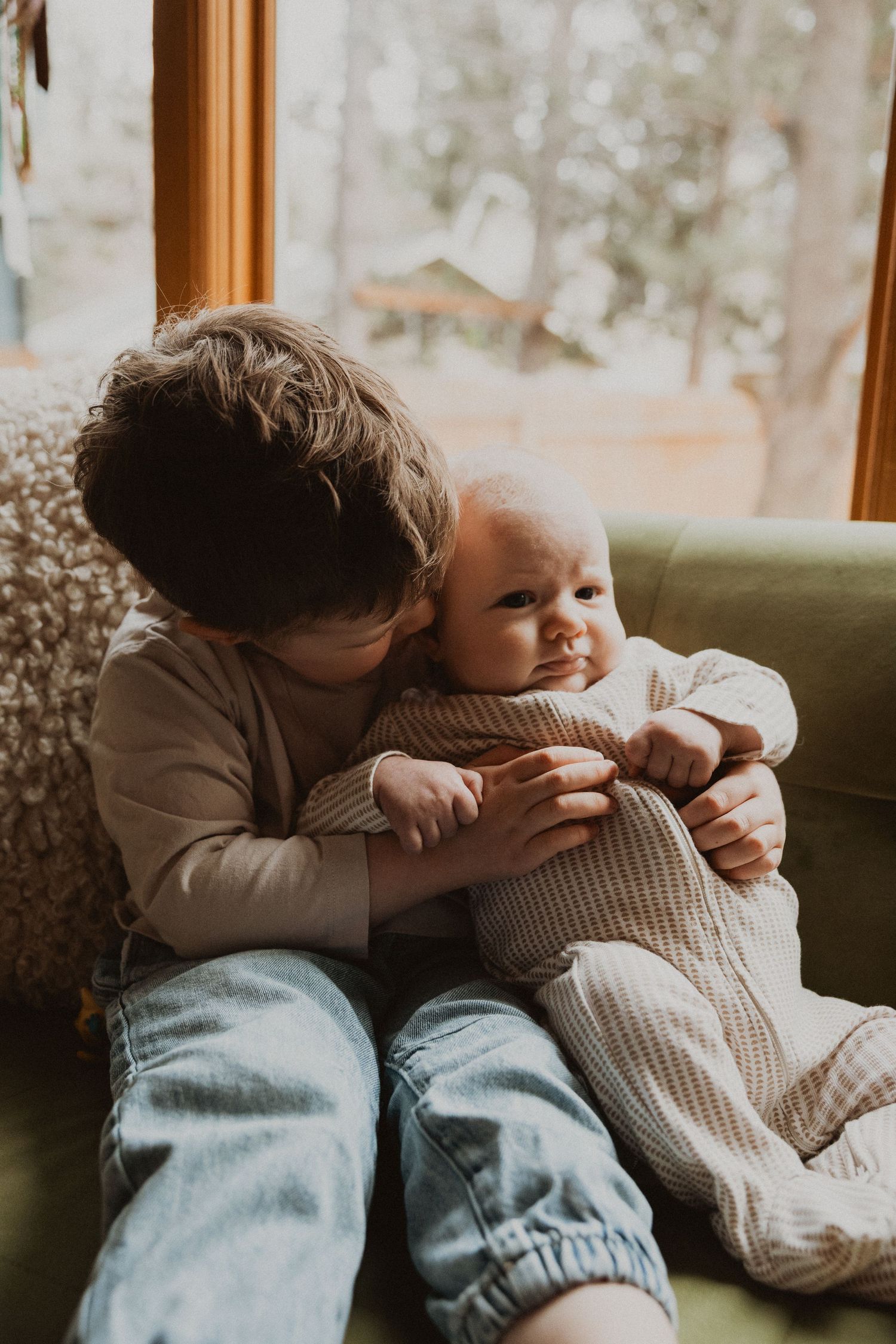 Young boy tenderly kissing newborn baby sibling while seated on green sofa by window.