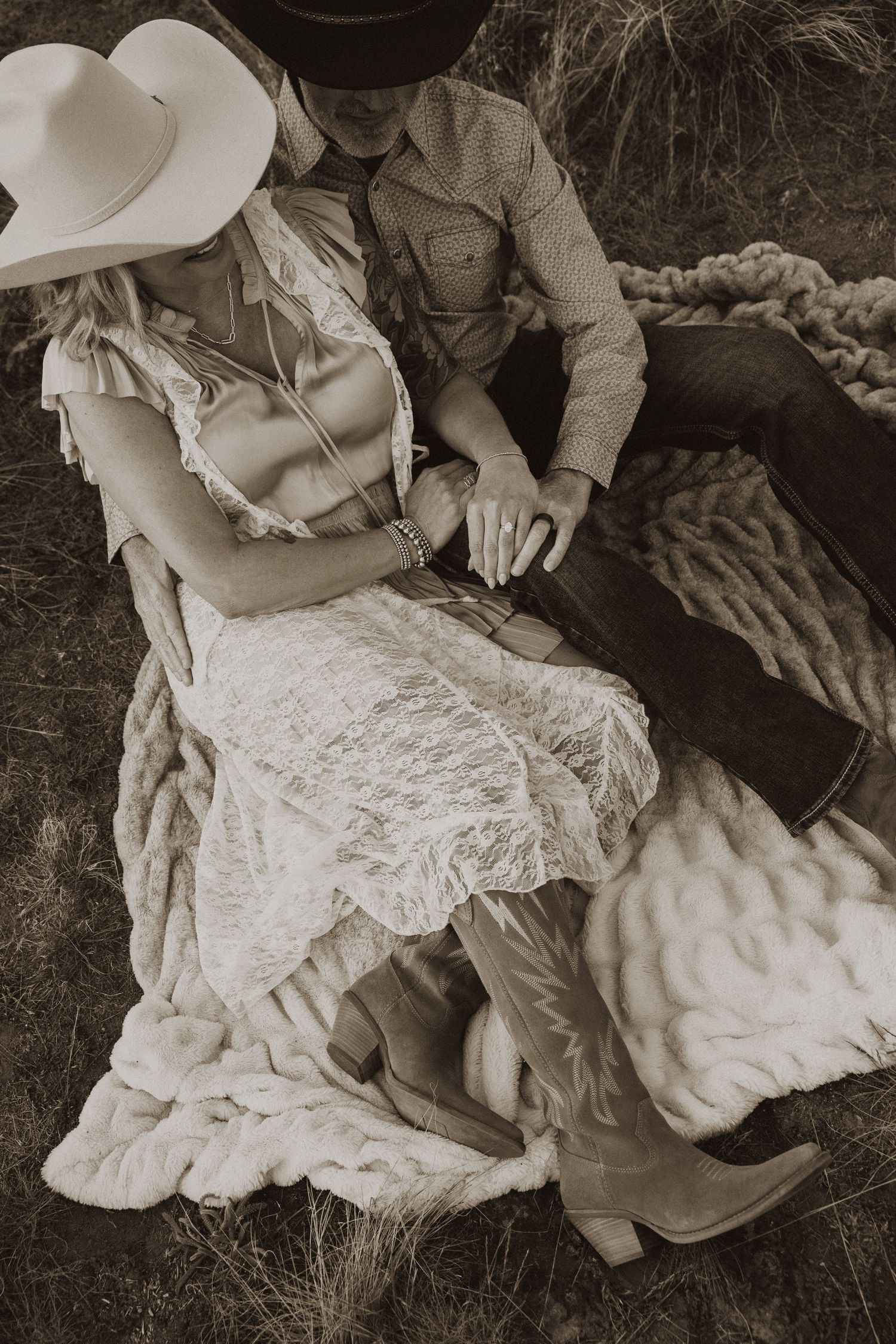Sepia-toned photo of a couple in western attire, cowboy hats, and boots relaxing on a fur blanket outdoors.