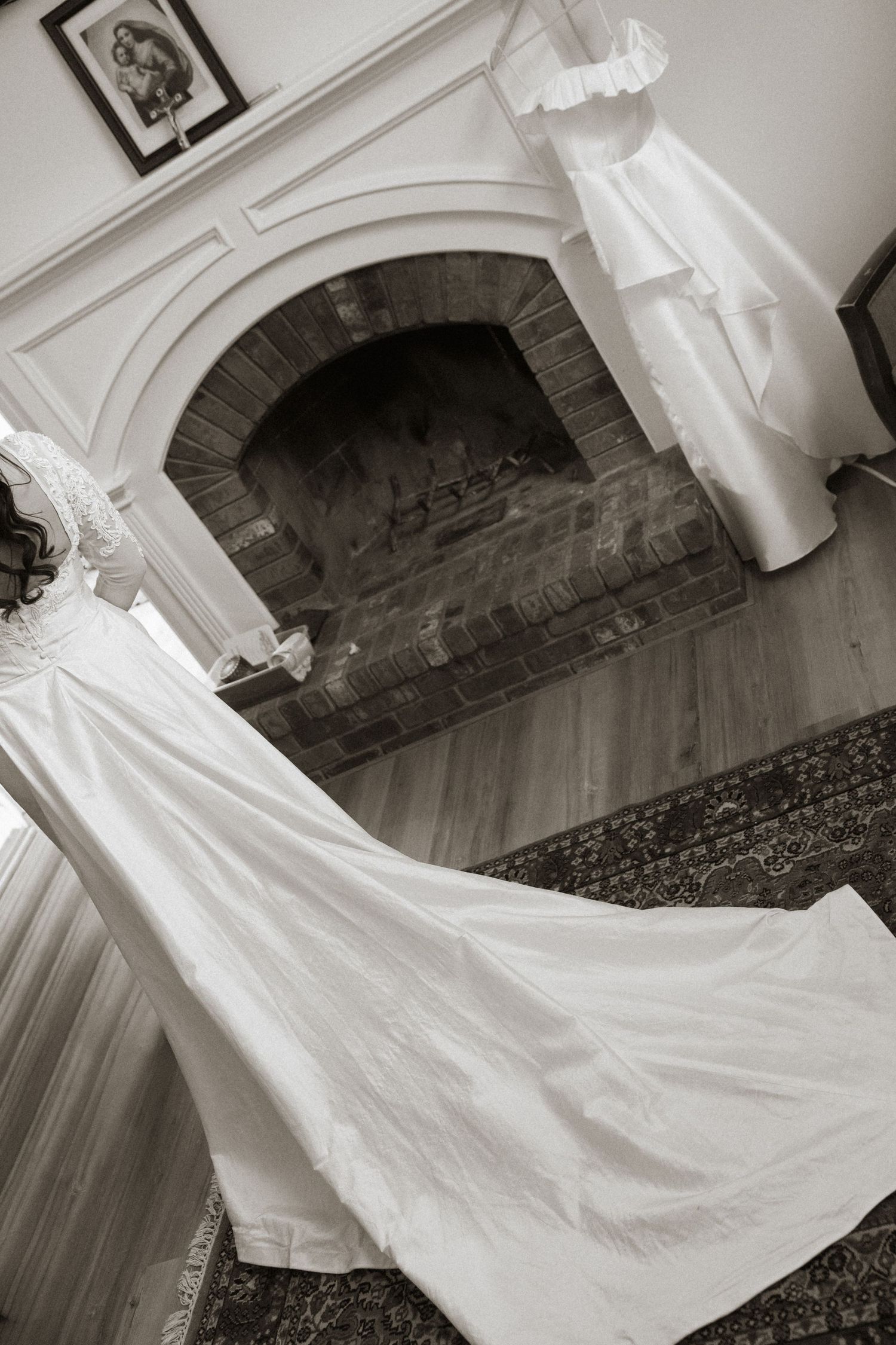 Black and white photo of a bridal gown with long train displayed near a rustic brick fireplace.