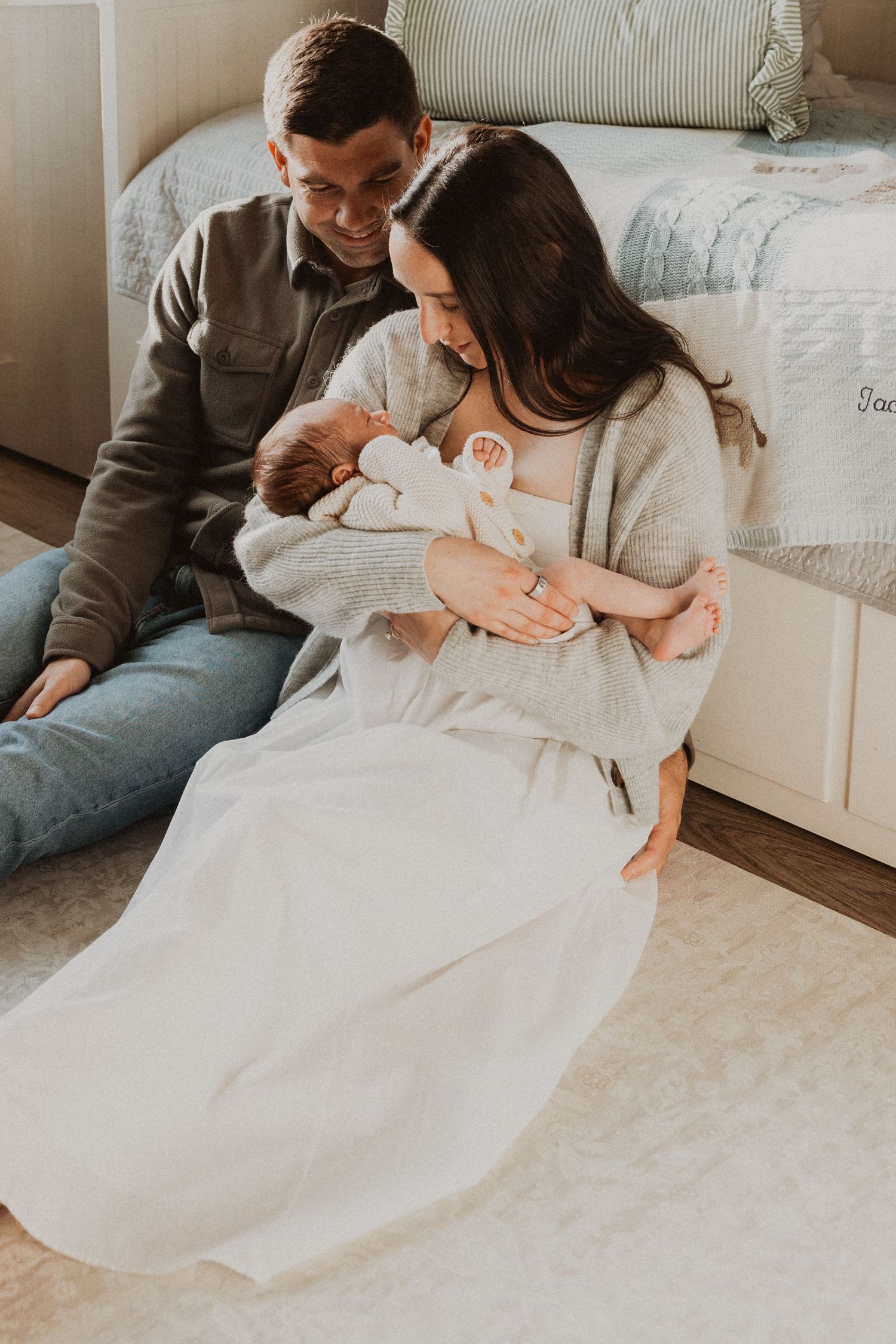 New parents sitting on floor holding newborn baby, mom in white dress and gray cardigan, dad in gray jacket smiling.