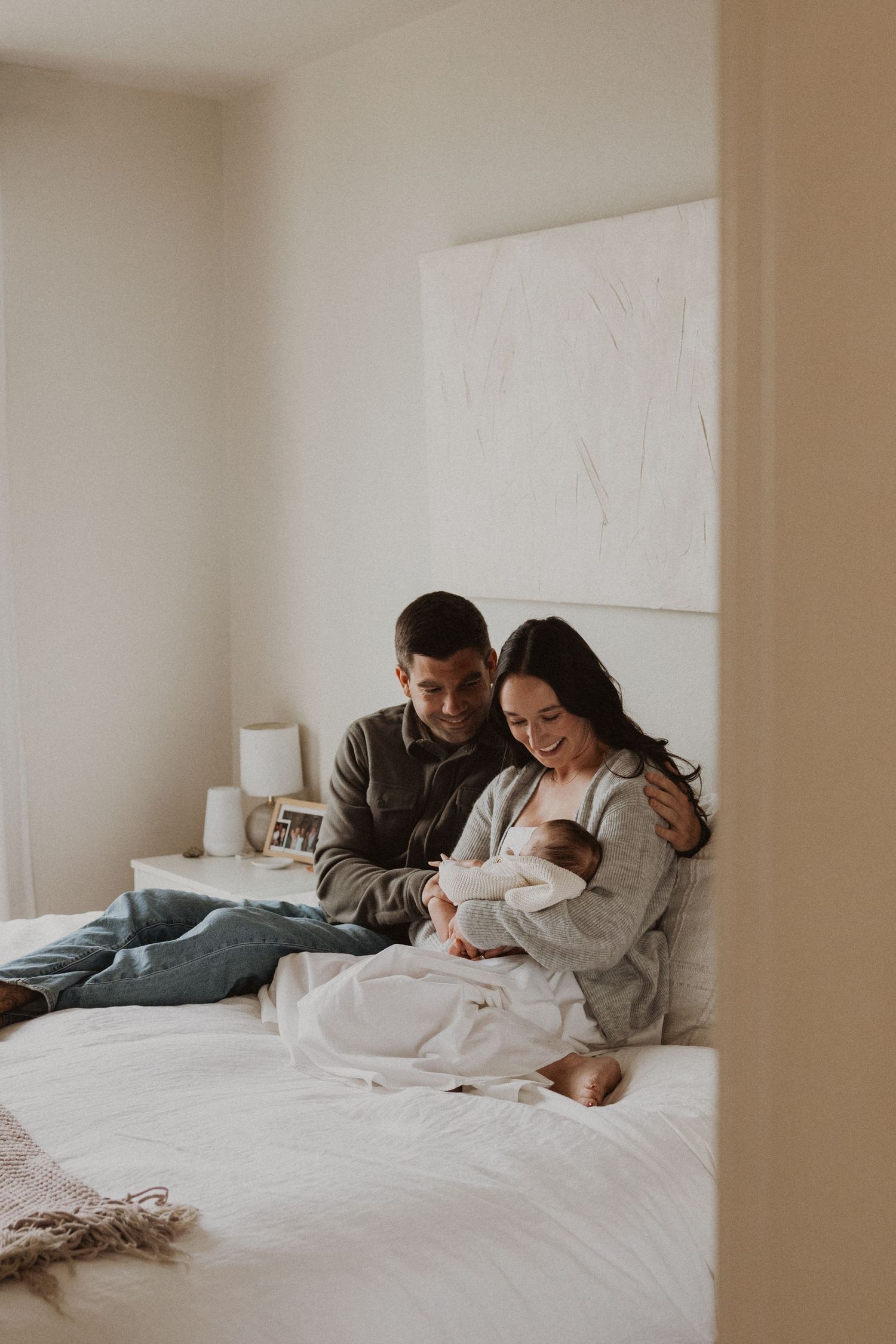 A couple sits on a white bed, smiling at their newborn baby wrapped in a blanket in a bright bedroom.