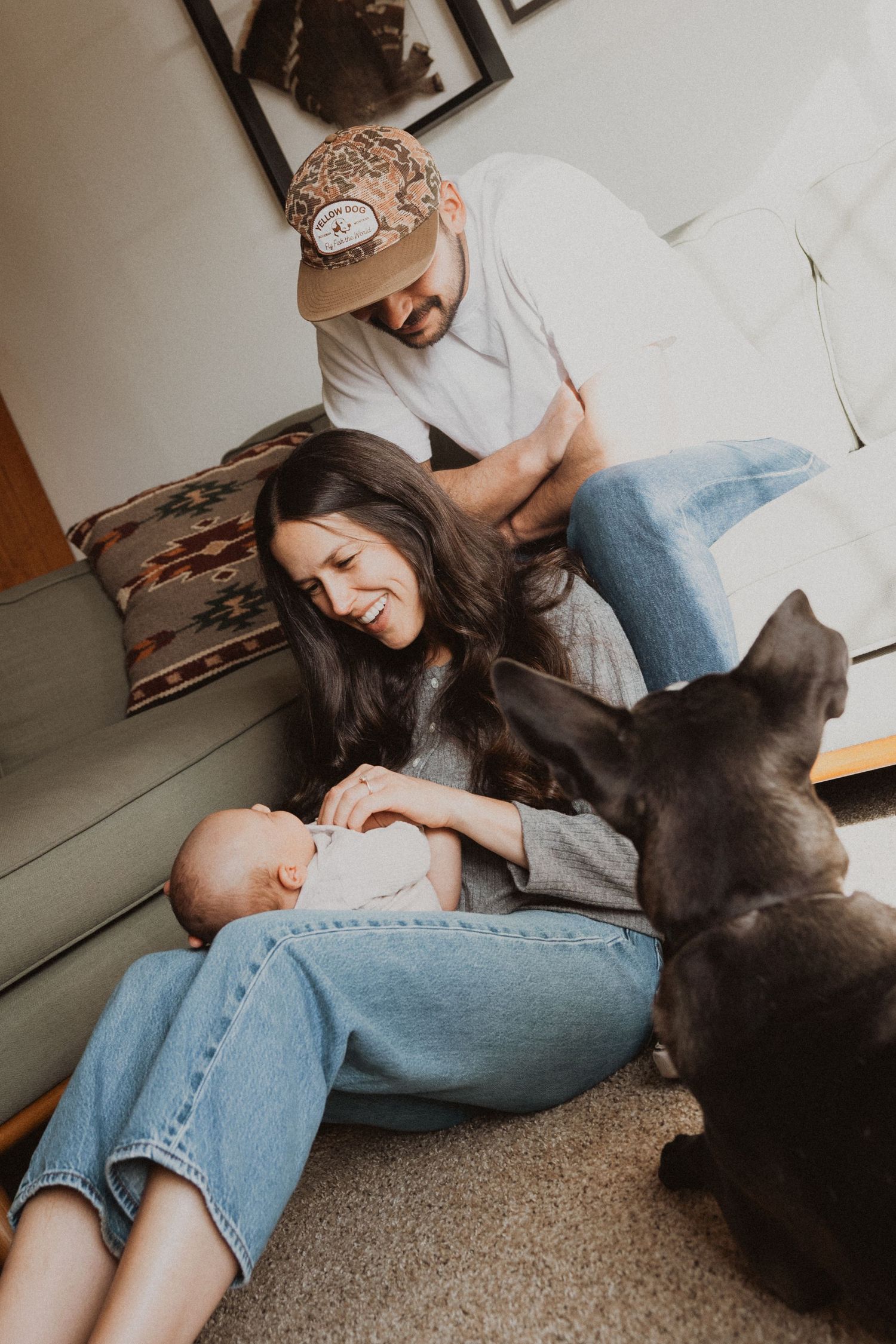 A smiling family with a newborn baby and a black dog relaxing together on the floor of a cozy living room.