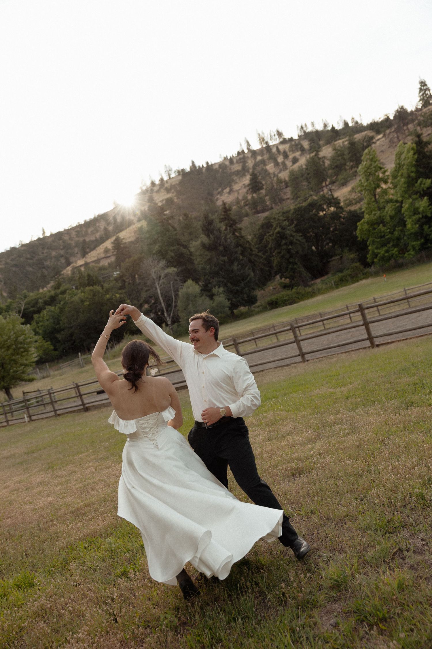 Bride and groom dancing and twirling in a sunlit field with mountains in the background.