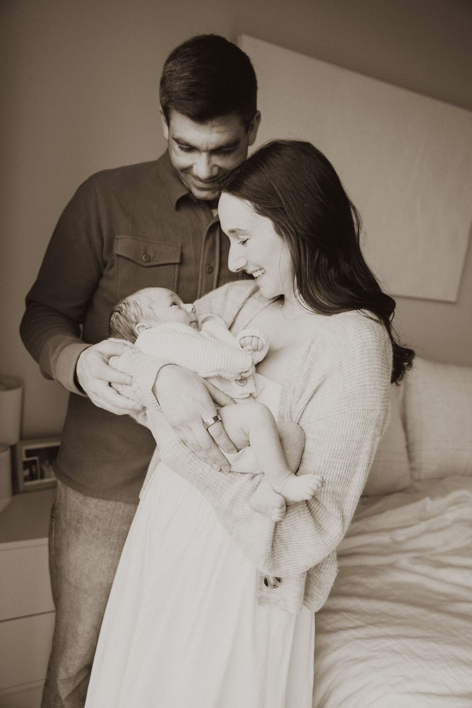 Sepia-toned photo of happy parents lovingly holding their newborn baby in a cozy bedroom setting.