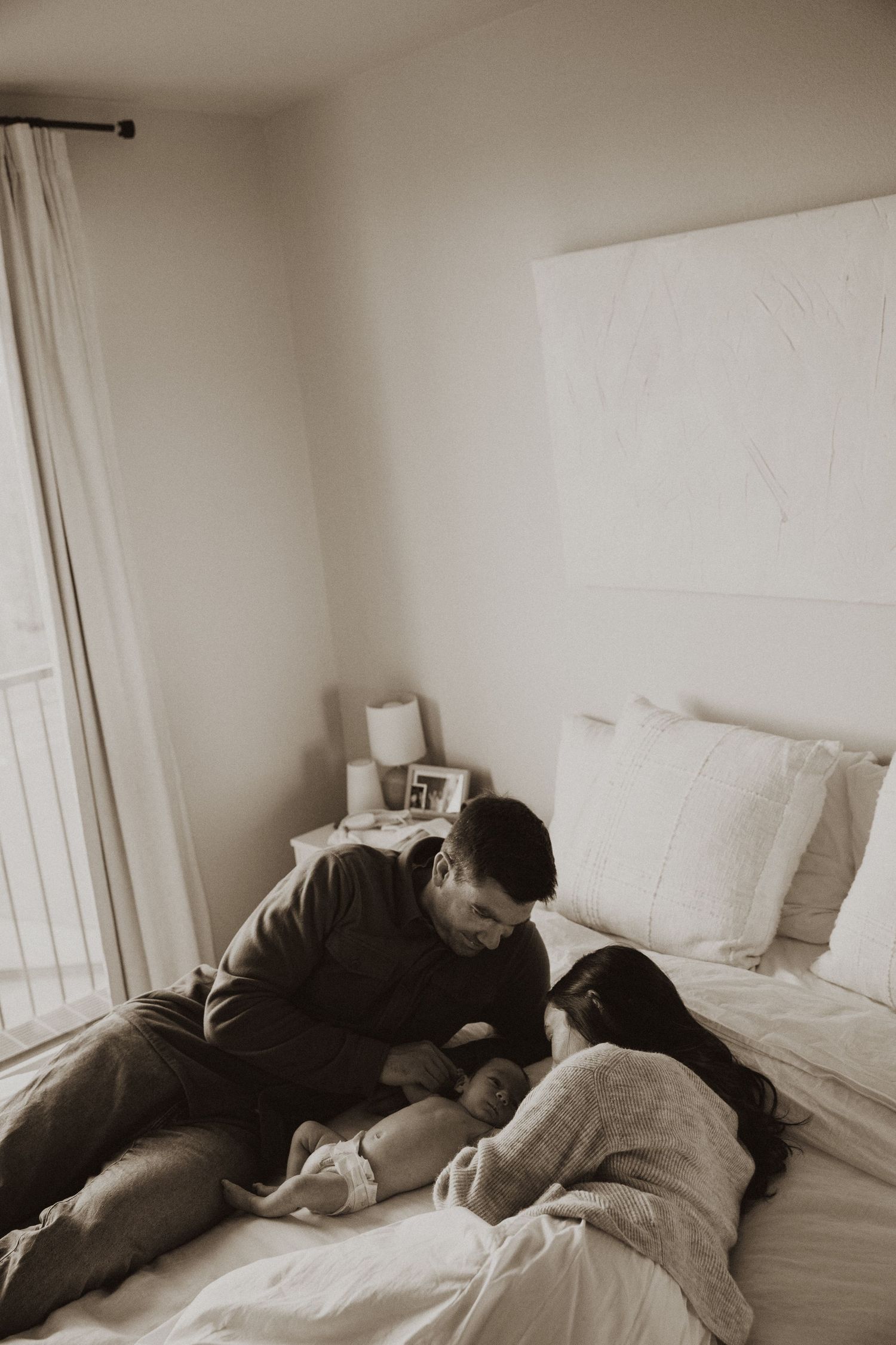 Sepia-toned photo of parents lying on bed together, lovingly gazing at their newborn baby in a cozy bedroom.