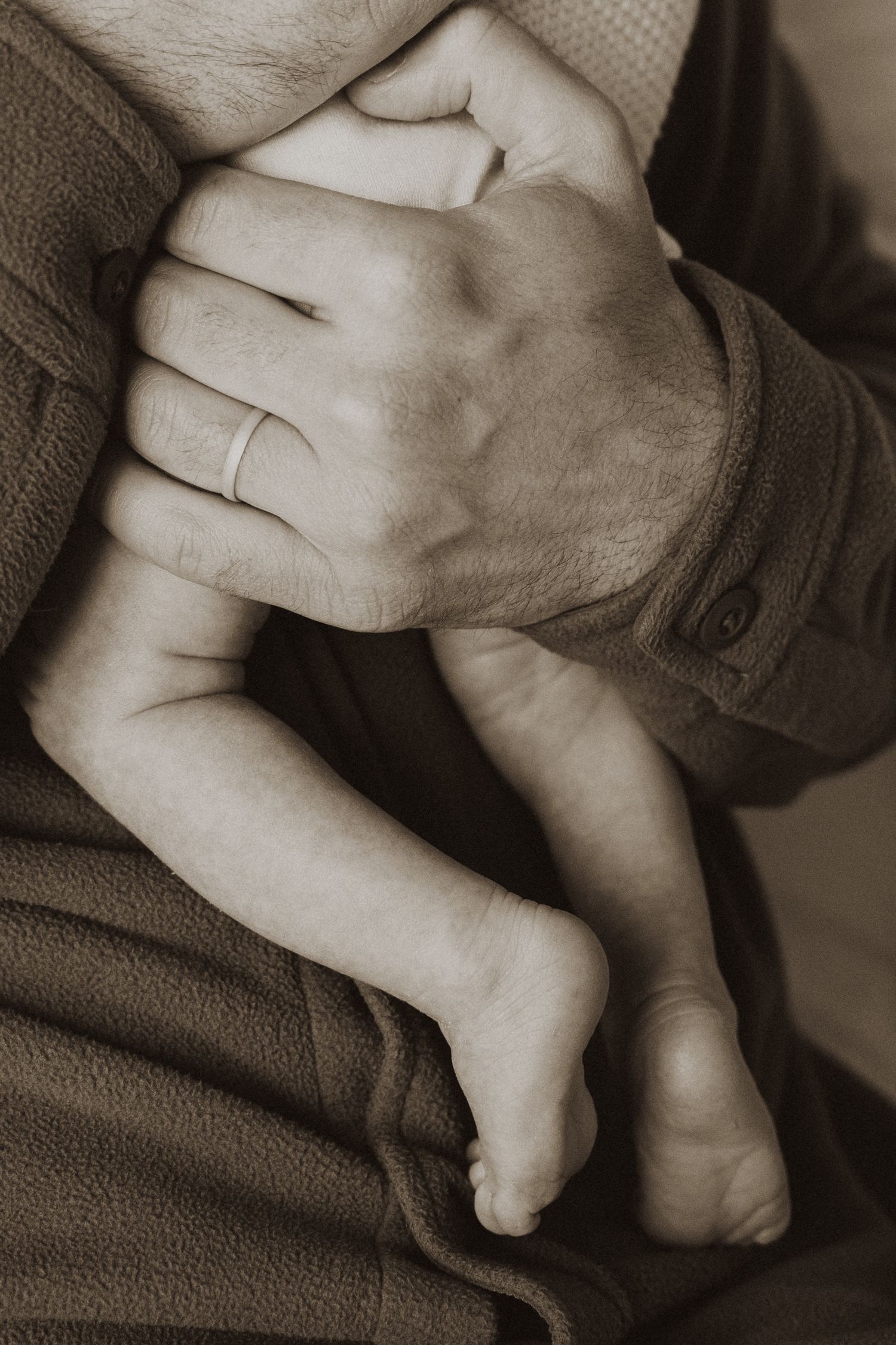 A parent's hand with wedding ring cradling a newborn baby's tiny feet and legs in a sepia-toned photo.