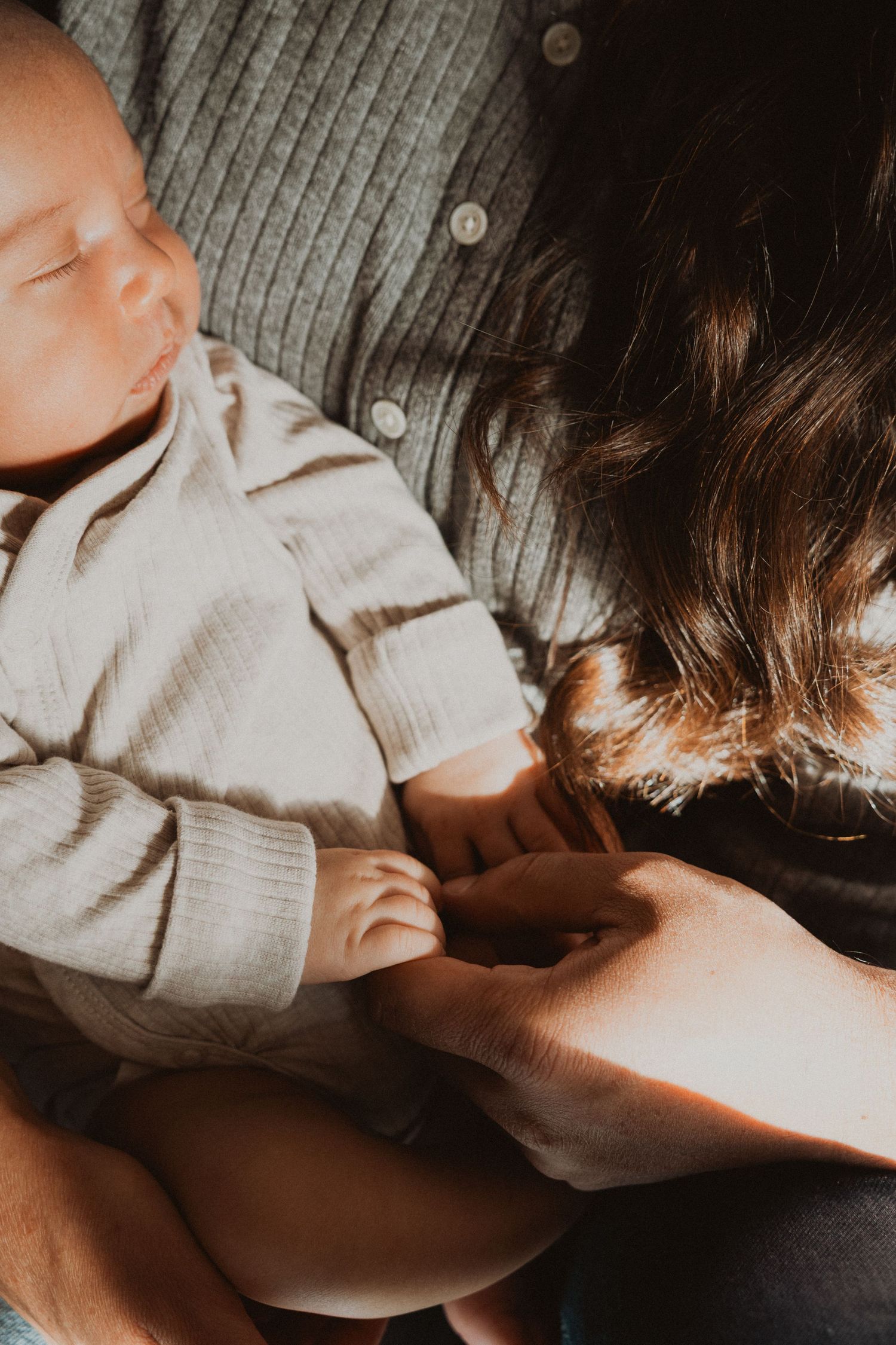 Newborn baby sleeping peacefully in parent's arms, wearing a soft gray onesie in warm natural light.