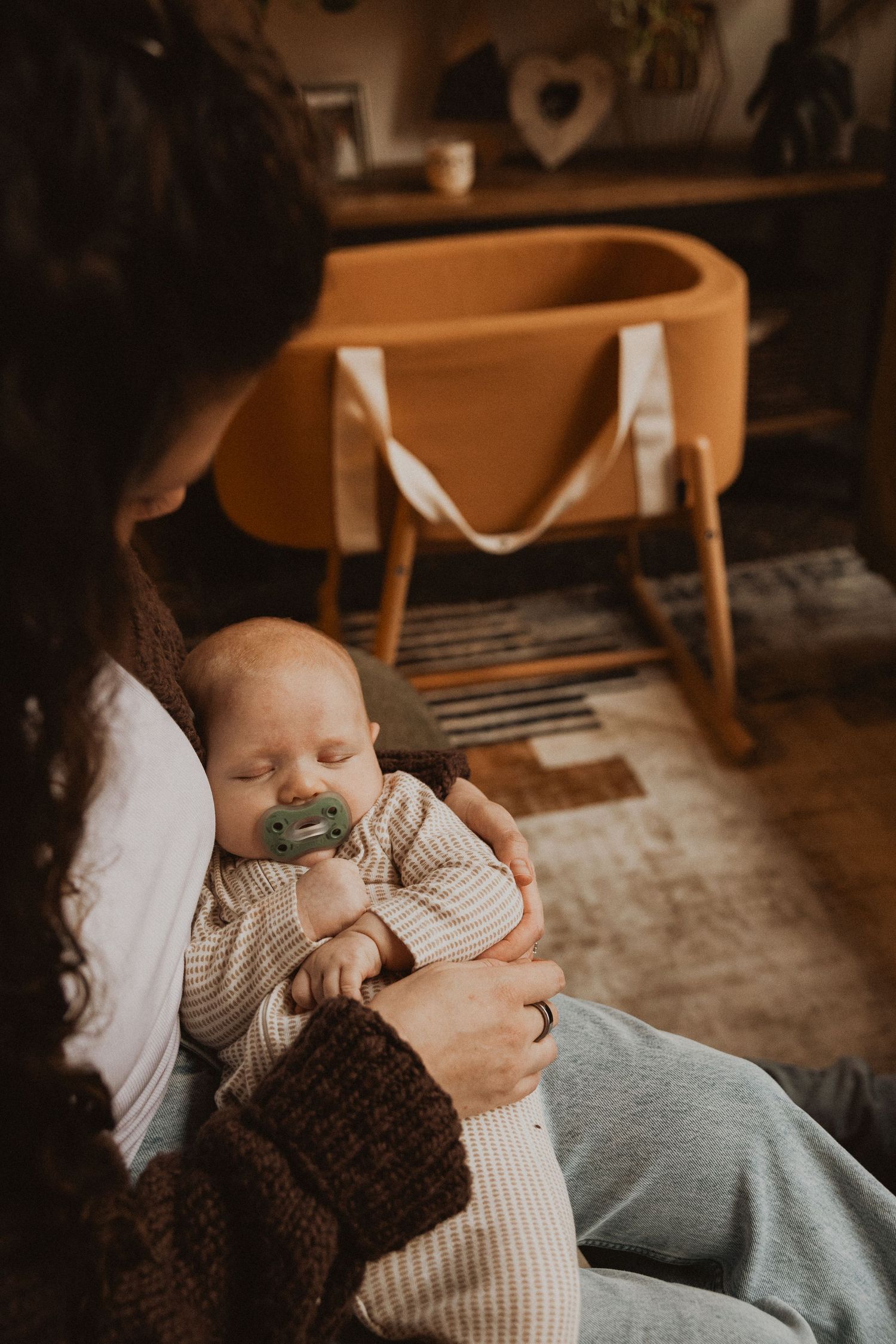 Mother cradling sleeping newborn with pacifier near a mustard yellow bassinet in cozy nursery.