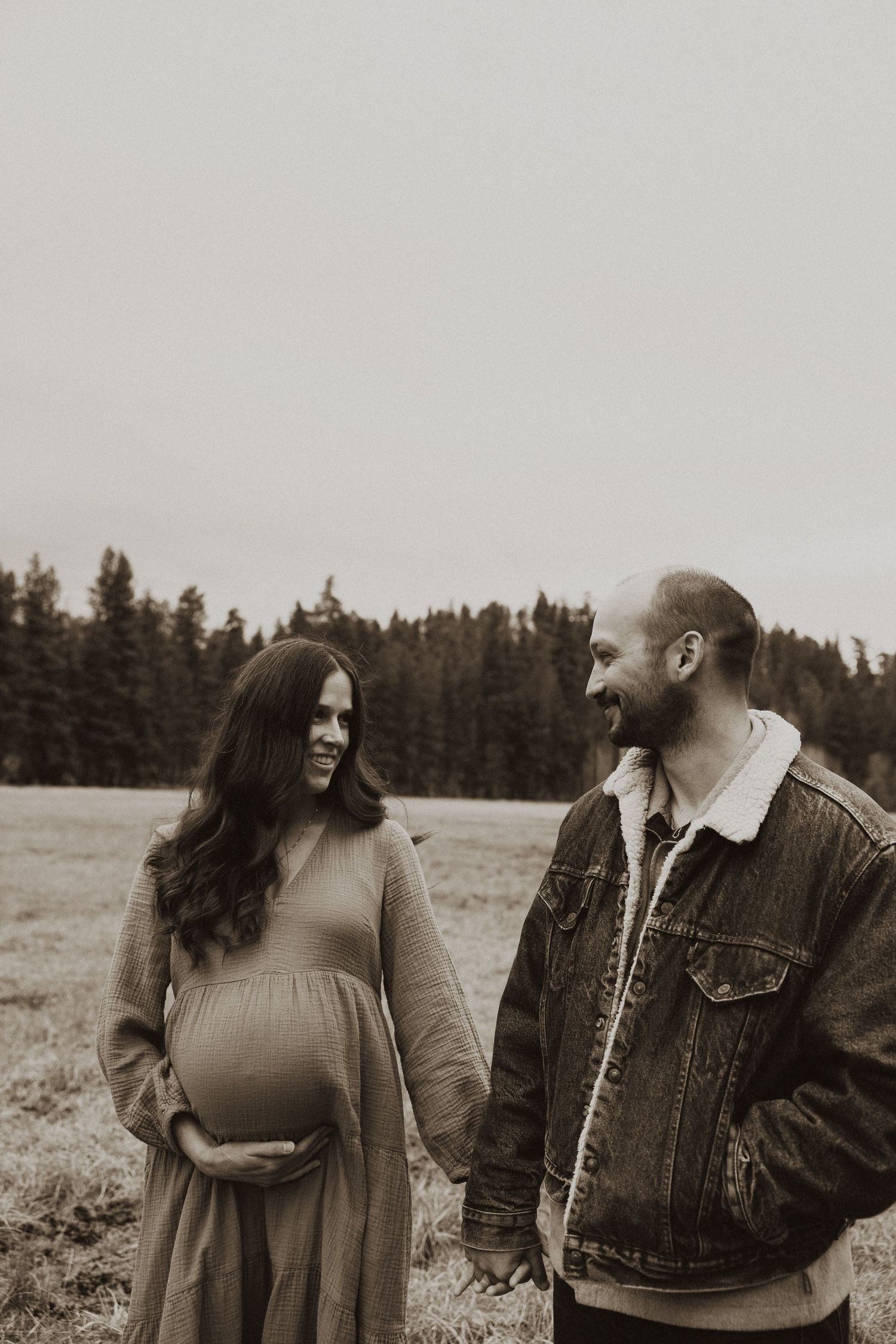 Smiling pregnant woman in maxi dress holds baby bump while holding hands with bearded partner in field.