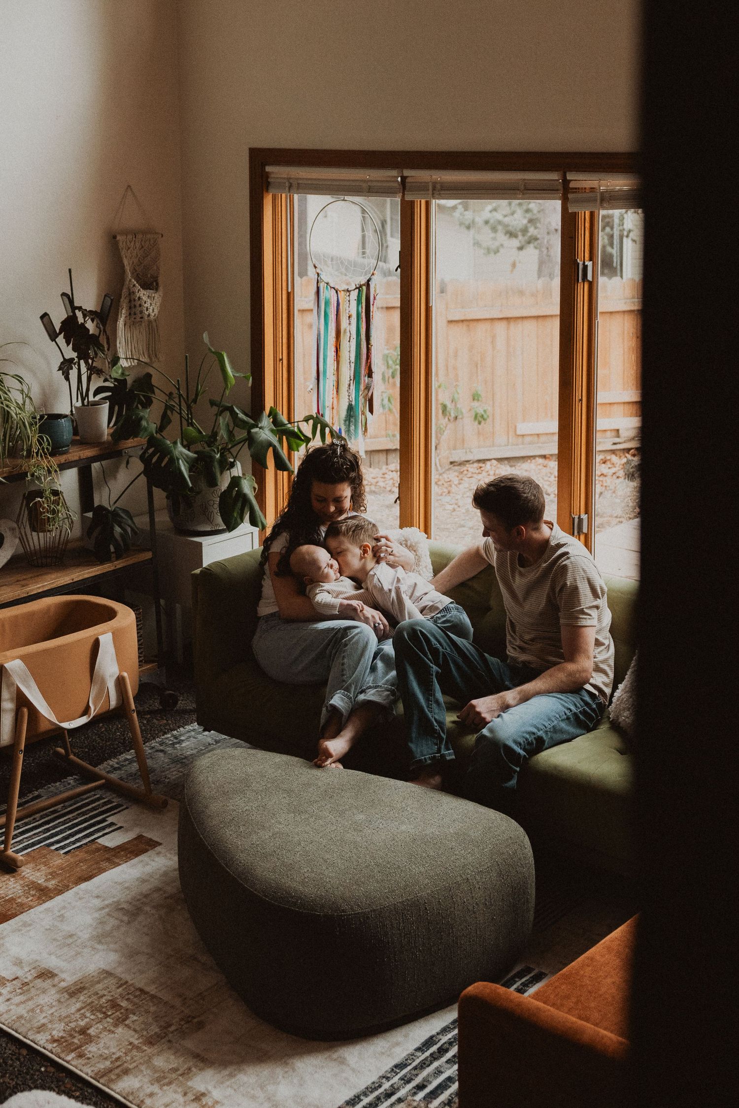 Family of four with newborn baby gathered together on green couch near sunny window.