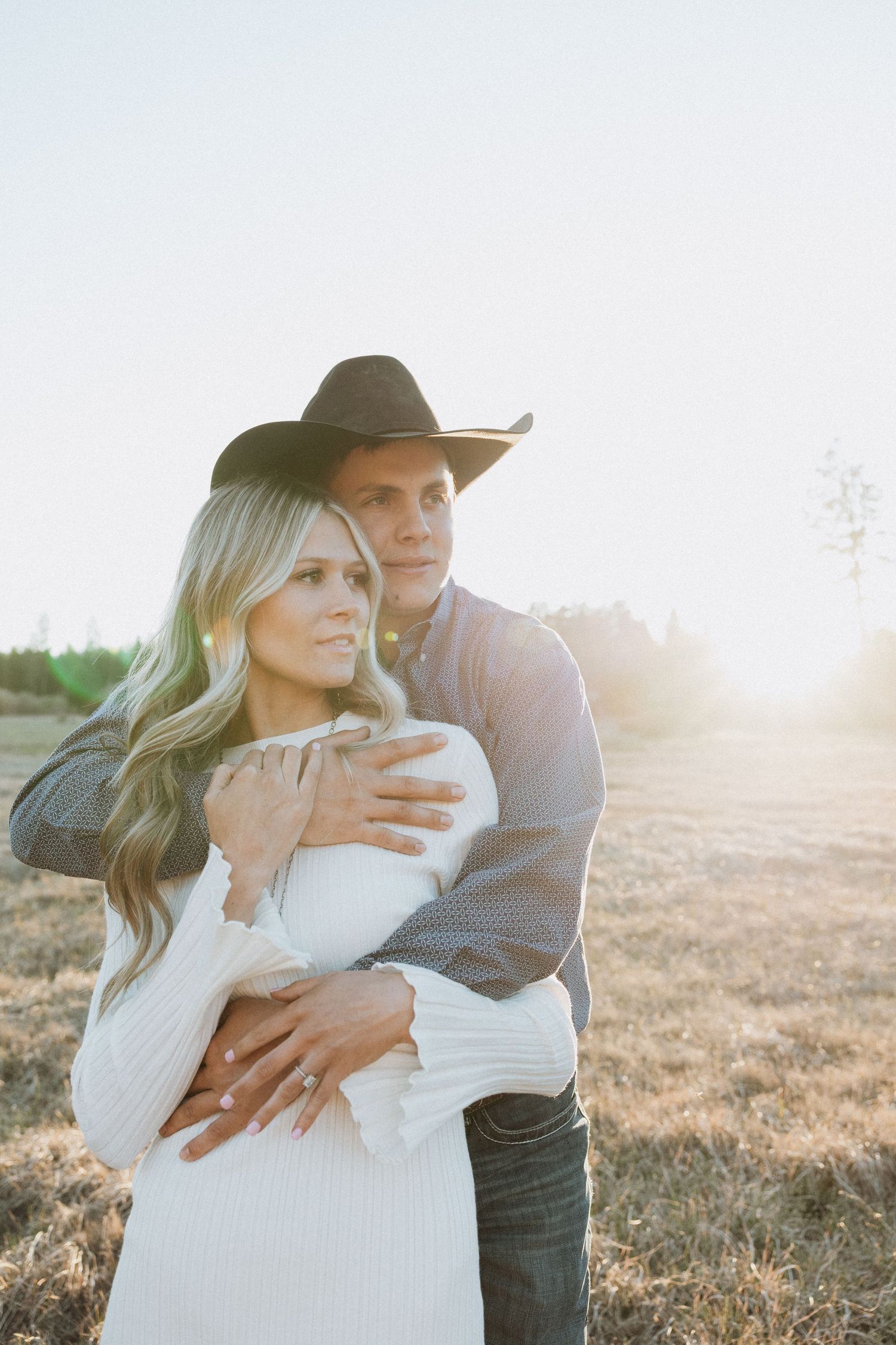 Couple embracing in golden sunlit field, man in black cowboy hat hugging woman in white dress outdoors.