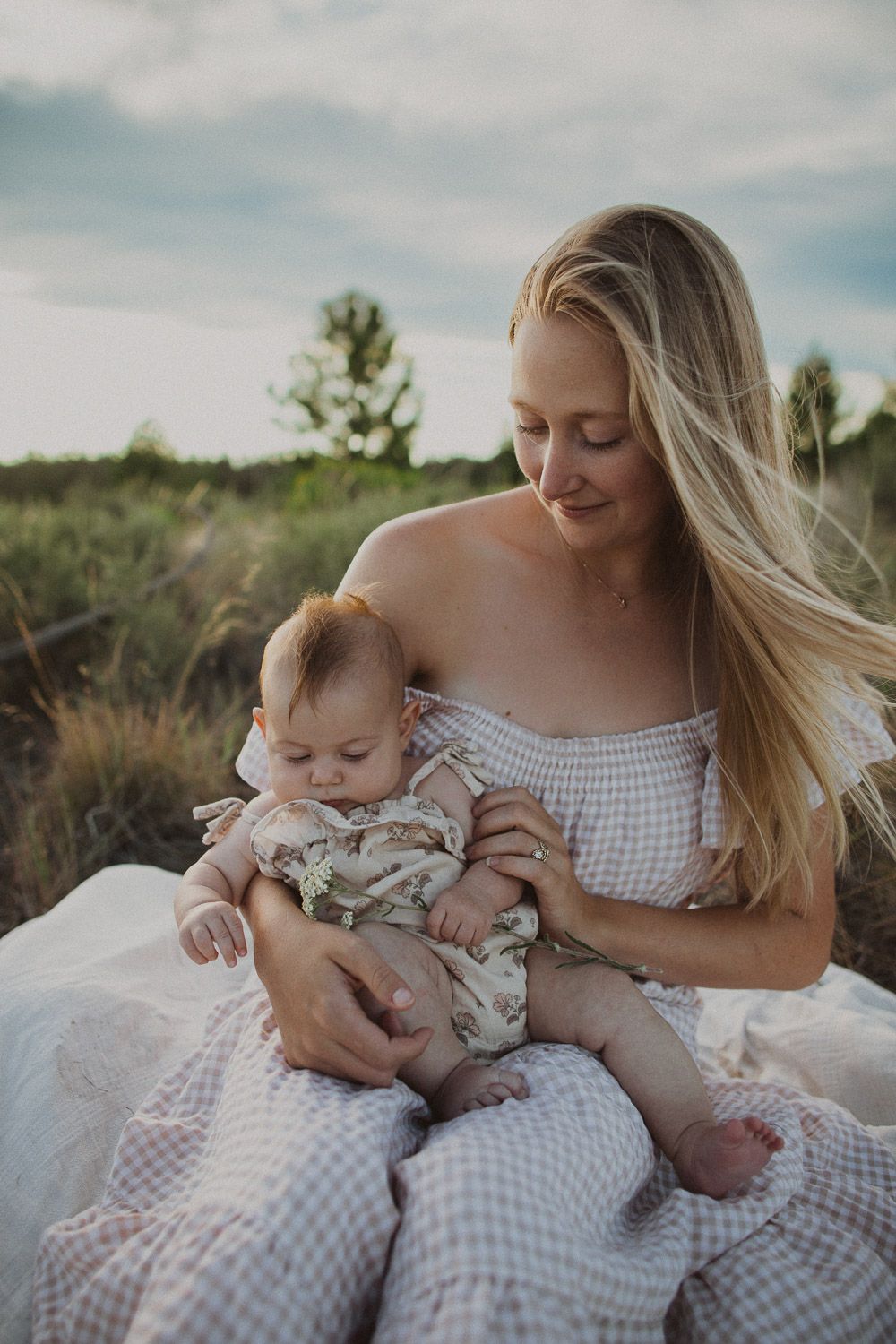 Intimate outdoor portrait of parent cradling baby while sitting on checkered blanket in grassy field.