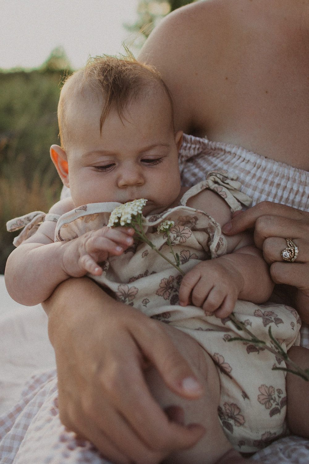 Close-up portrait of baby in floral outfit examining small white flowers outdoors.