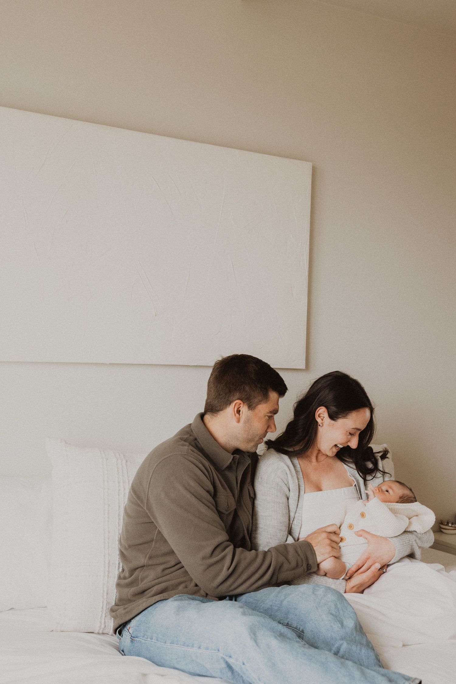 A couple sits on a white bed, lovingly gazing at their newborn baby in a soft, neutral-toned bedroom.