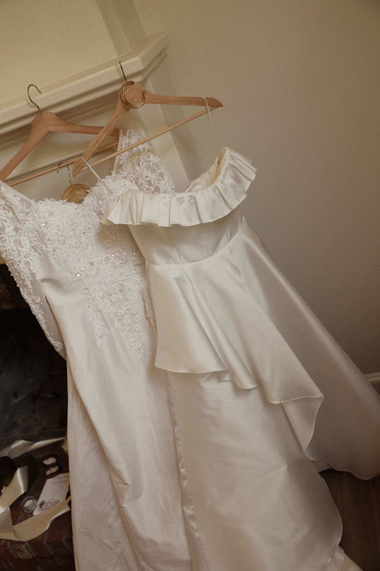 Two elegant white wedding dresses hanging on wooden hangers, one lace and one satin with ruffled neckline.