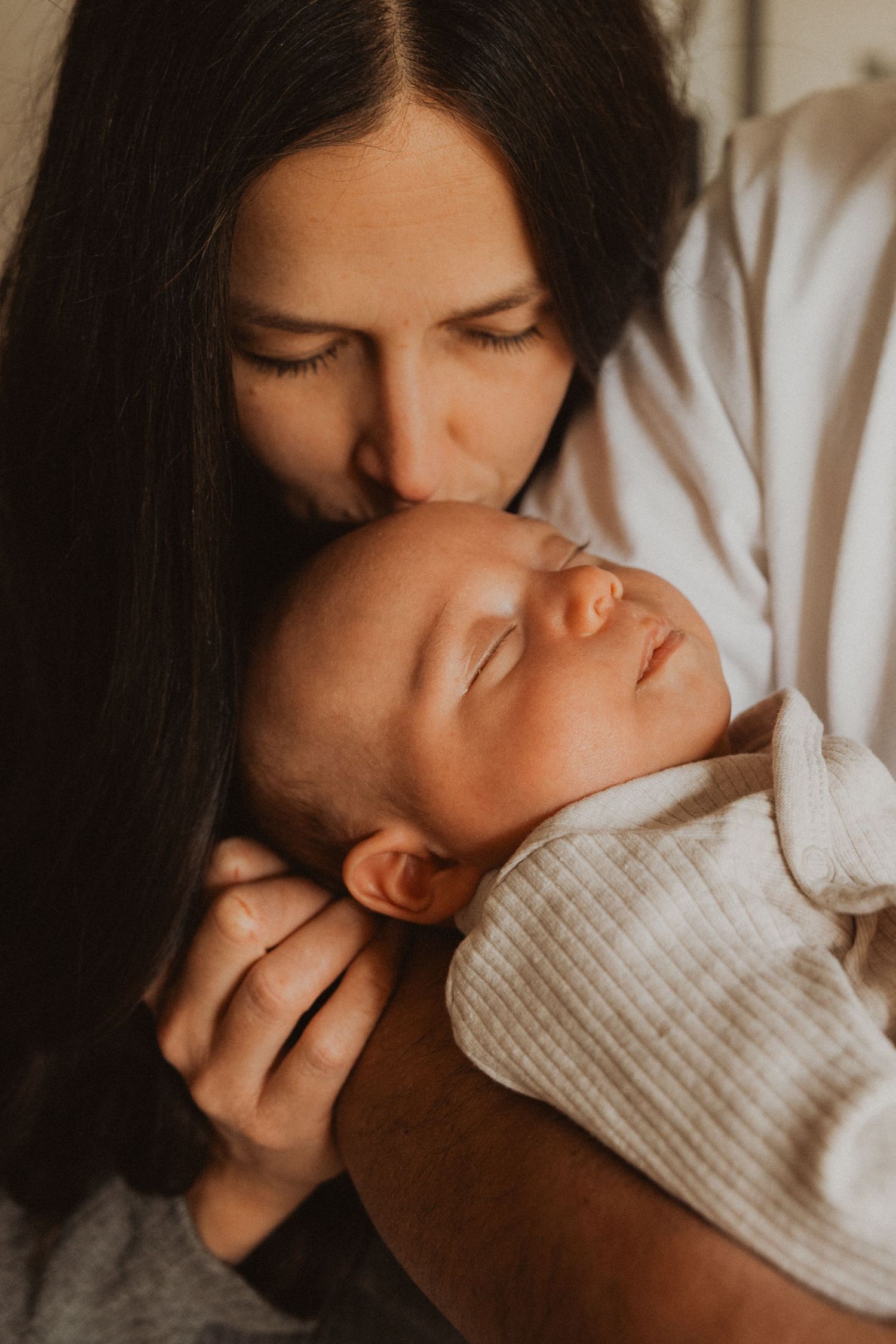 A dark-haired mother gently kisses her sleeping newborn baby wrapped in a soft white outfit.