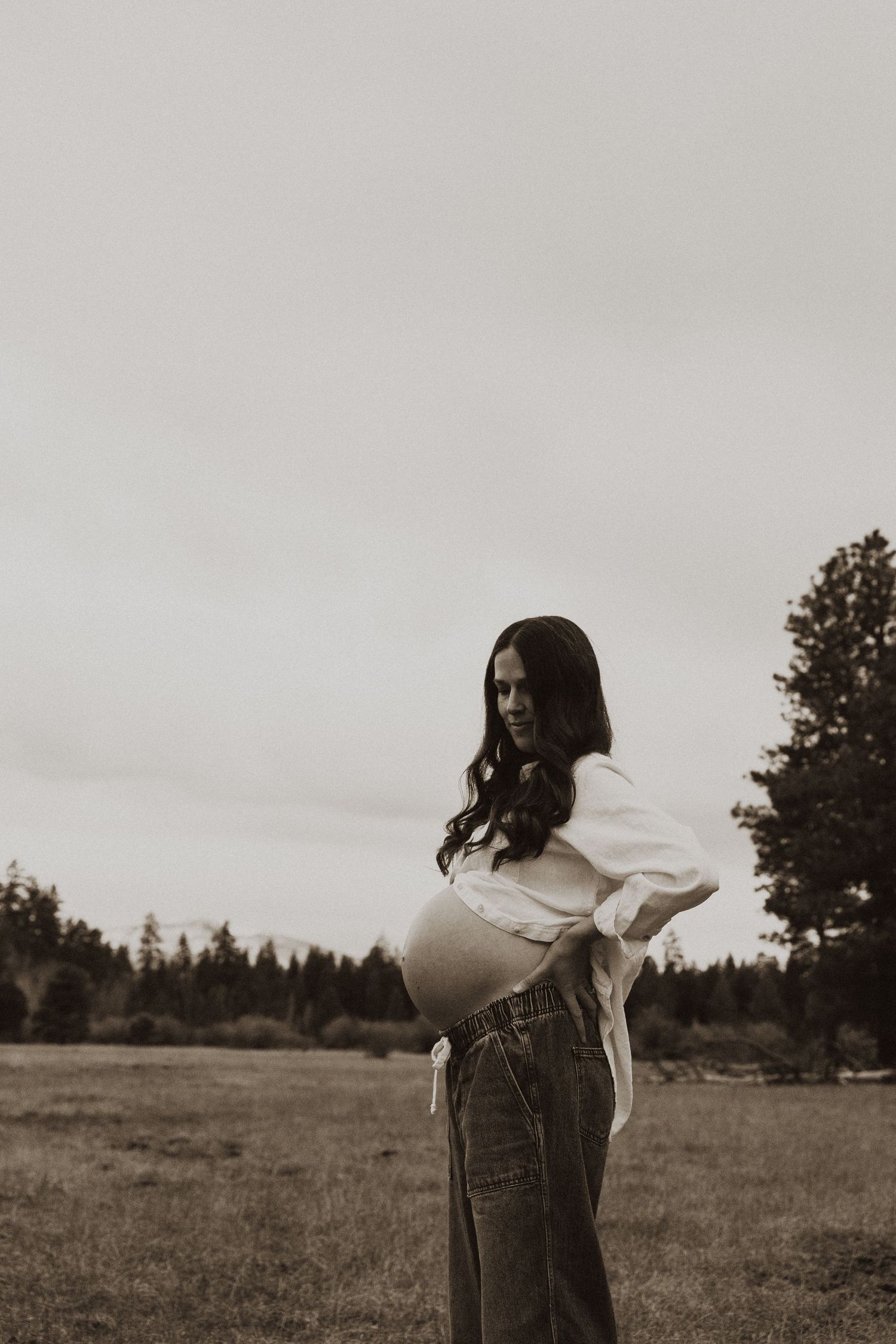 Black and white photo of a smiling pregnant woman in white top and jeans standing in an open field.