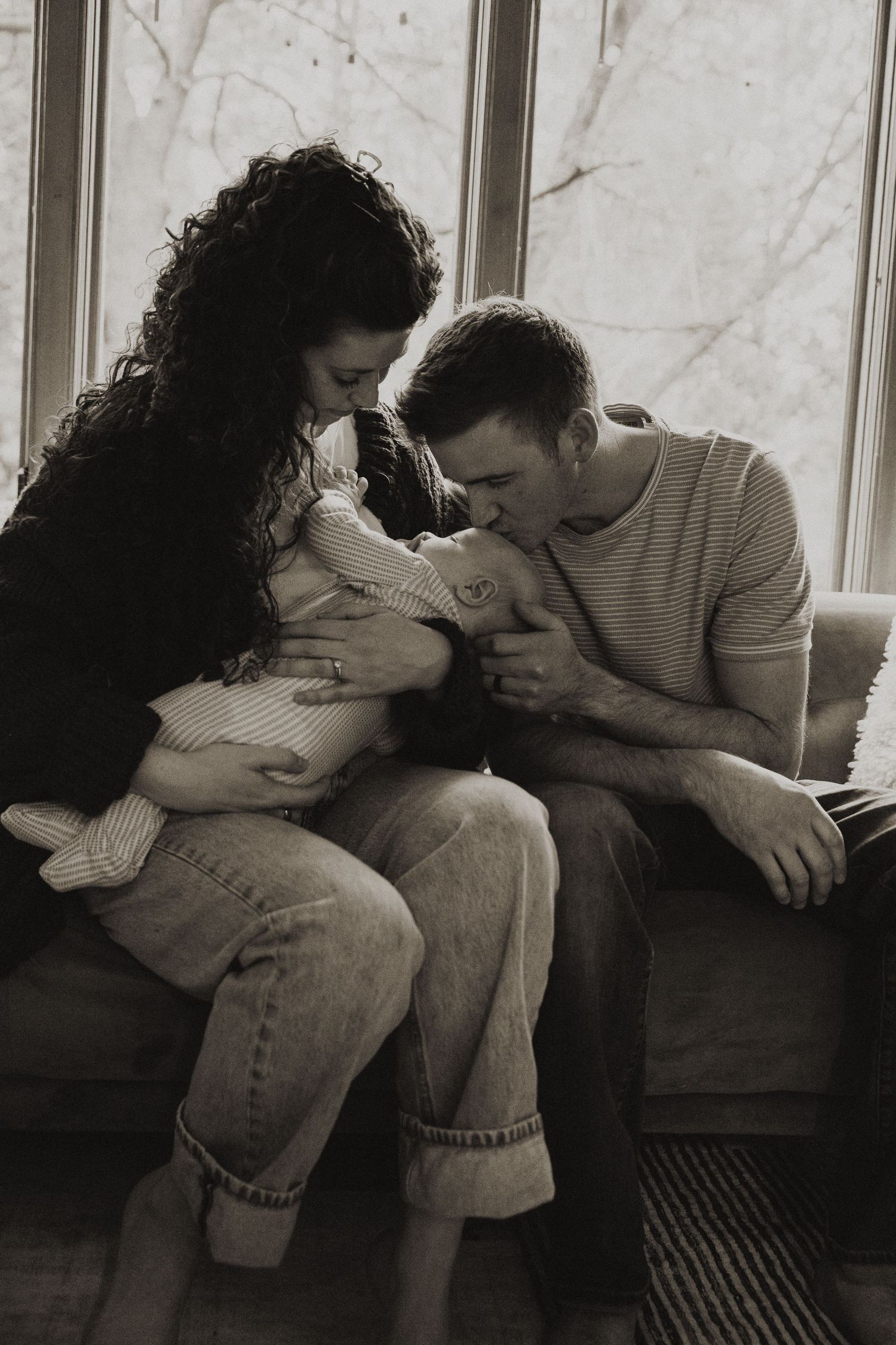New parents leaning in to kiss their newborn baby while seated by a large window.