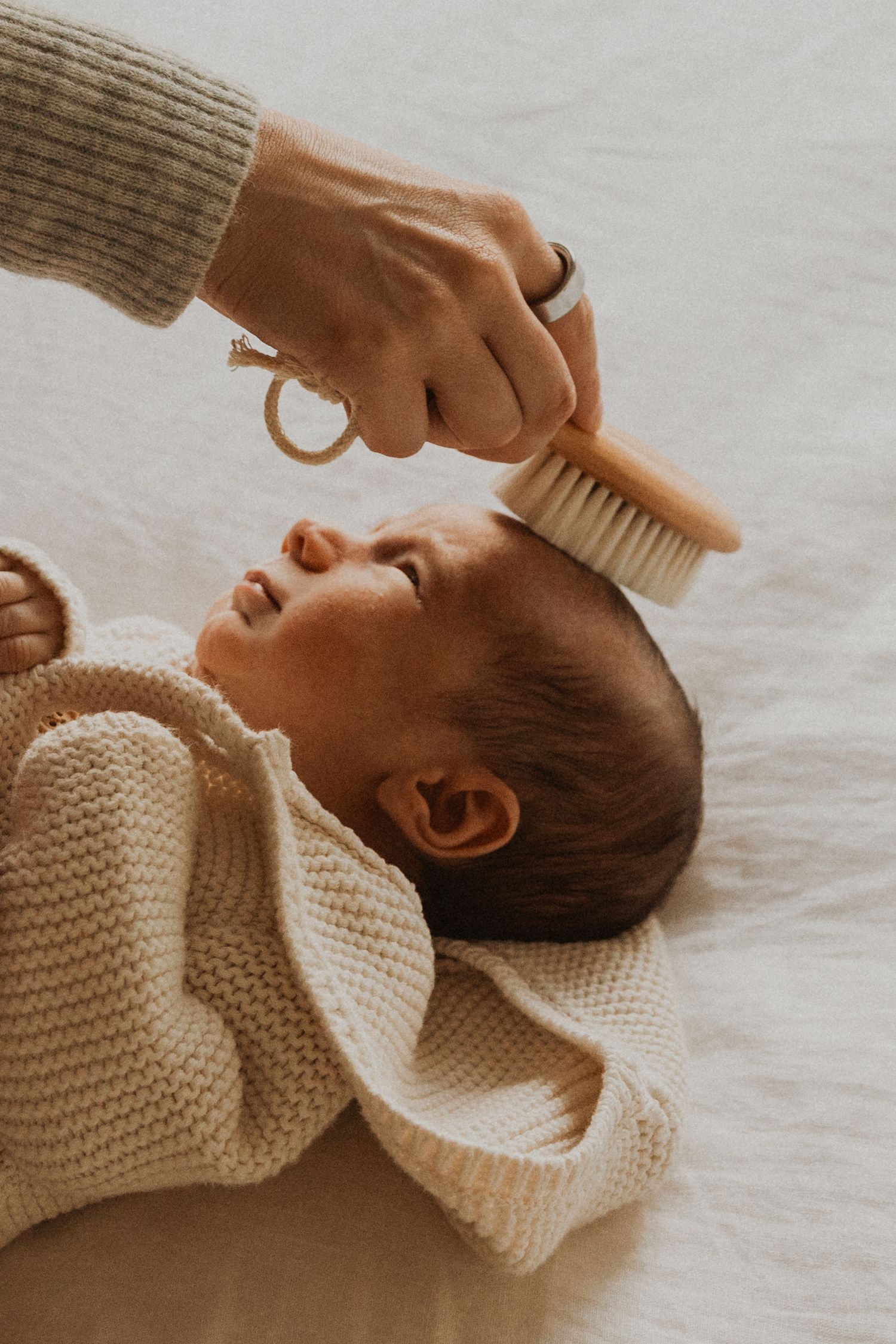 A gentle hand brushing a newborn baby's hair with a soft wooden brush while the infant lies on a white blanket.