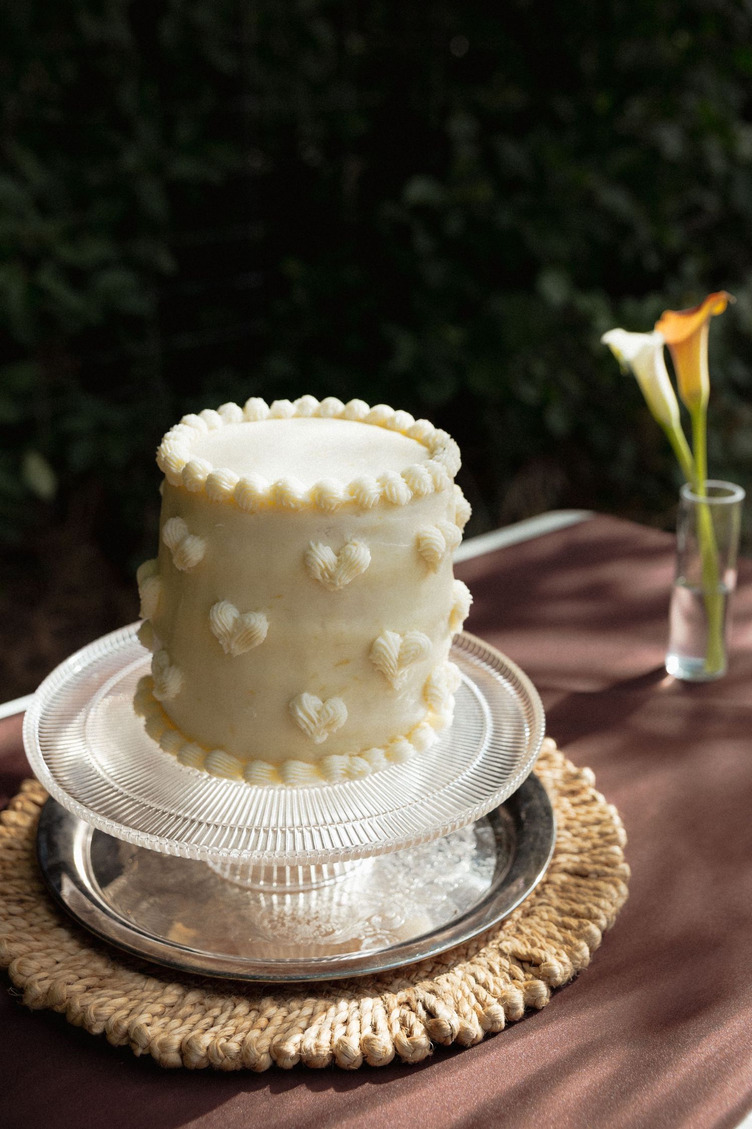 Elegant white wedding cake decorated with hearts on a silver stand at an outdoor garden reception.