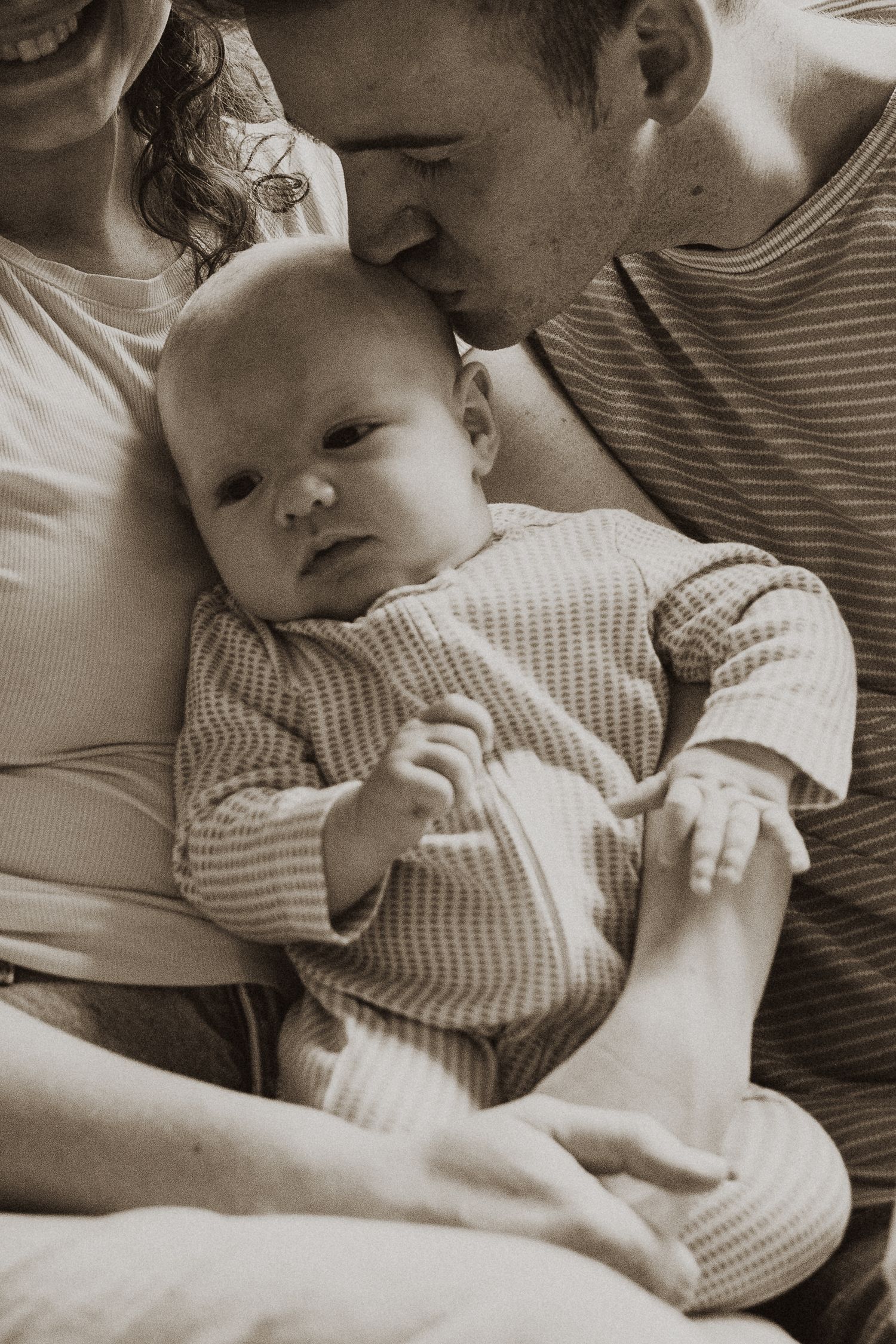 Close-up sepia photo of father gently kissing newborn baby held by mother on a sofa.