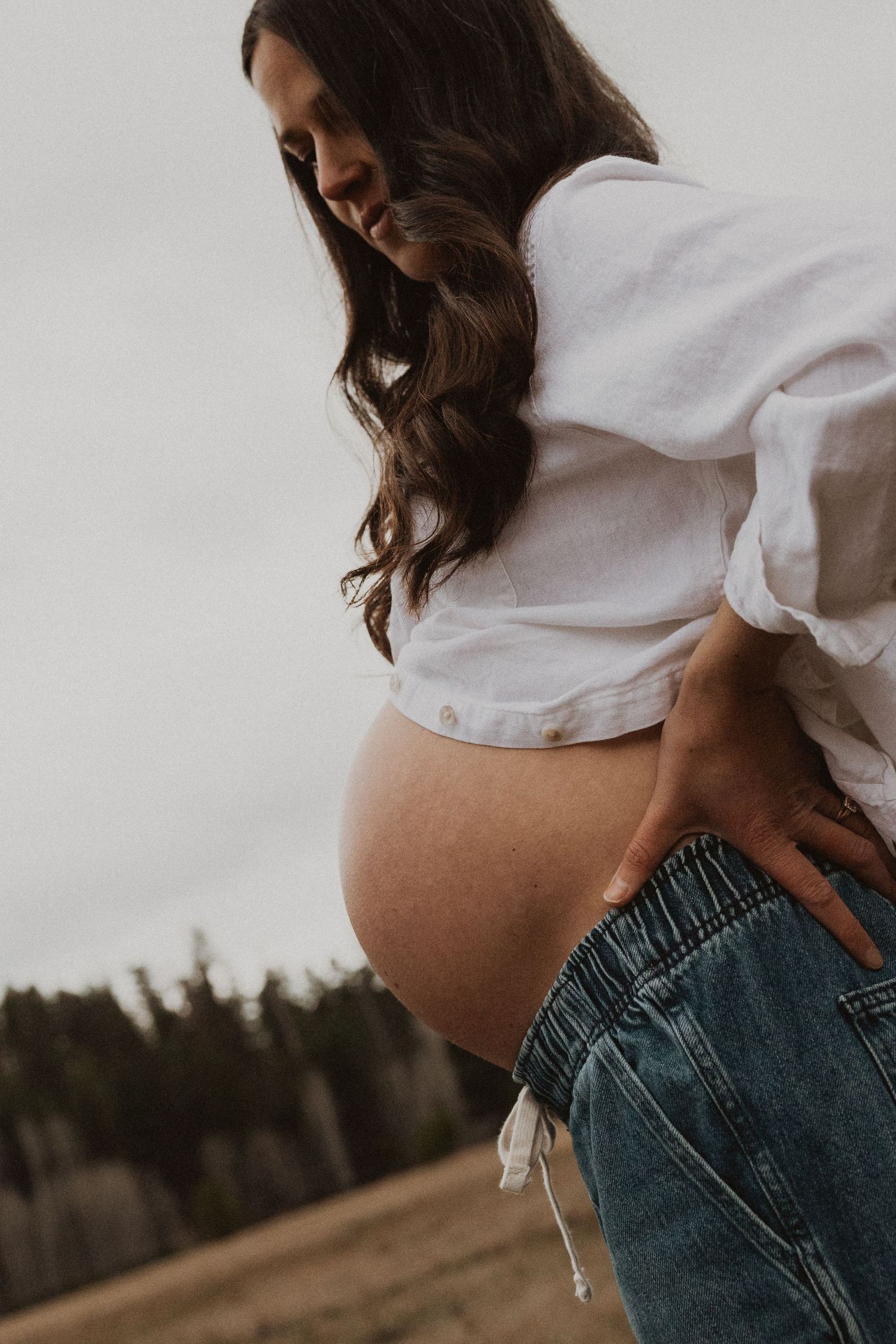 Pregnant woman in white blouse and jeans looks down at her bare baby bump outdoors in a field.