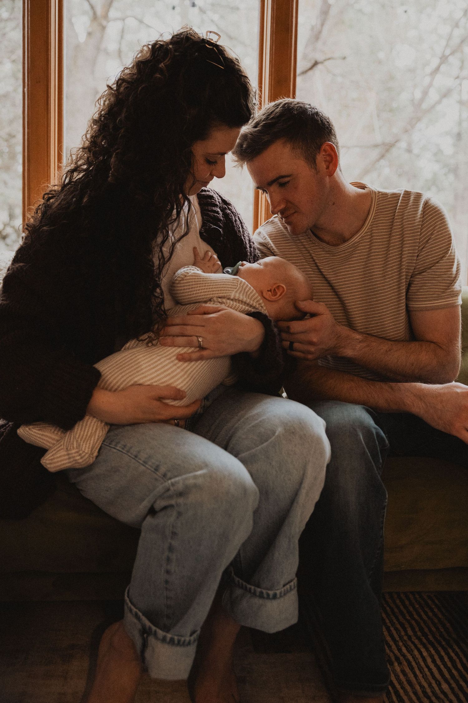 New parents sitting together by window smiling at their newborn baby in warm light.