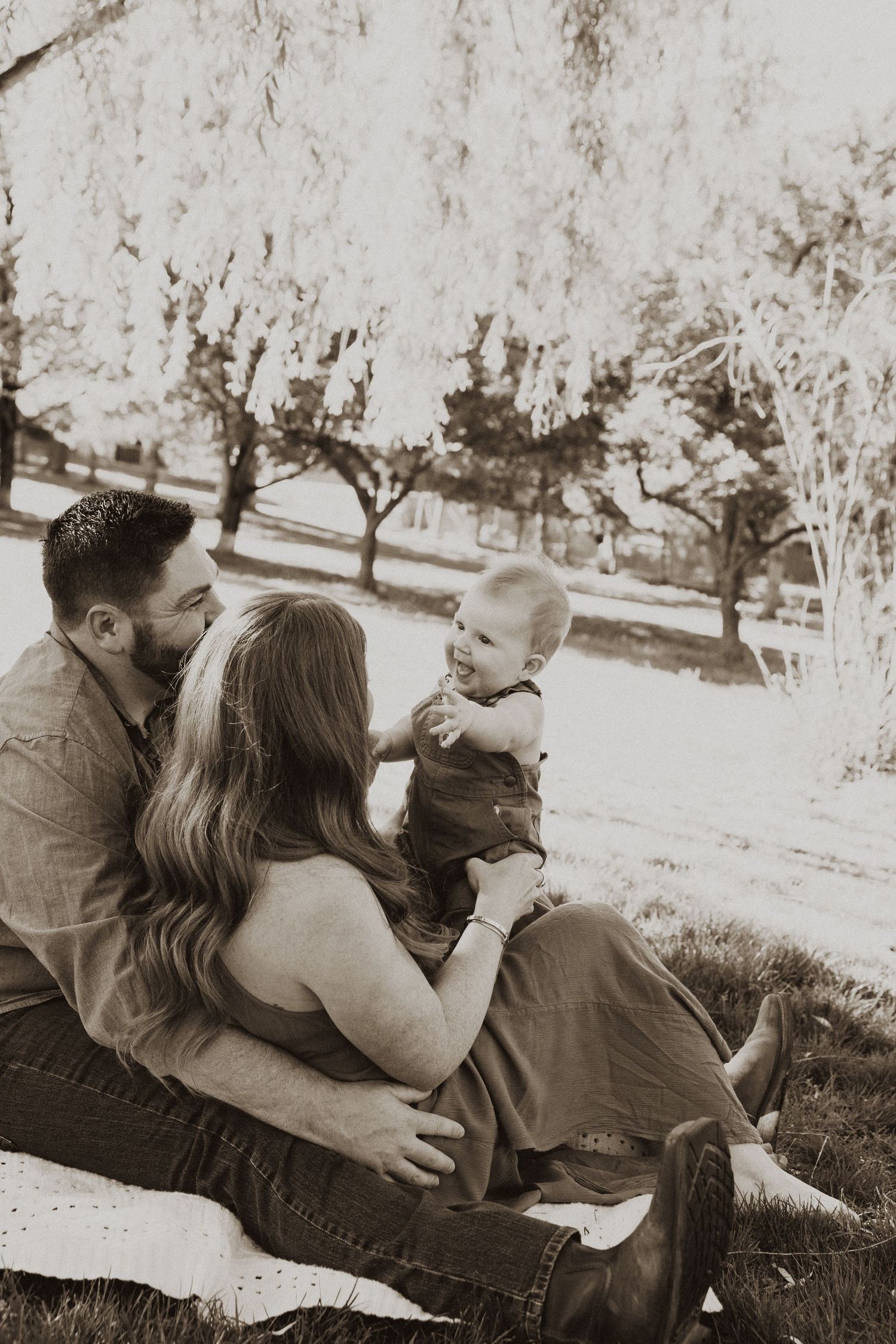 Sepia-toned family portrait of parents sitting outdoors on blanket with smiling baby in park setting.